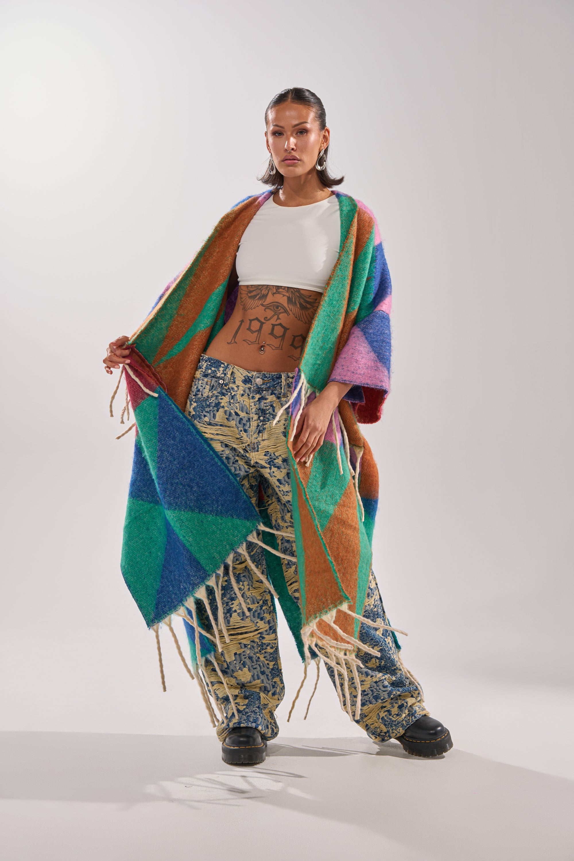 A person stands confidently against a plain light background, wearing the RENEWED DREAM TAPESTRY PANT—wide-leg, two-tone denim with a distressed pattern—paired with a white crop top and colorful geometric-patterned shawl.