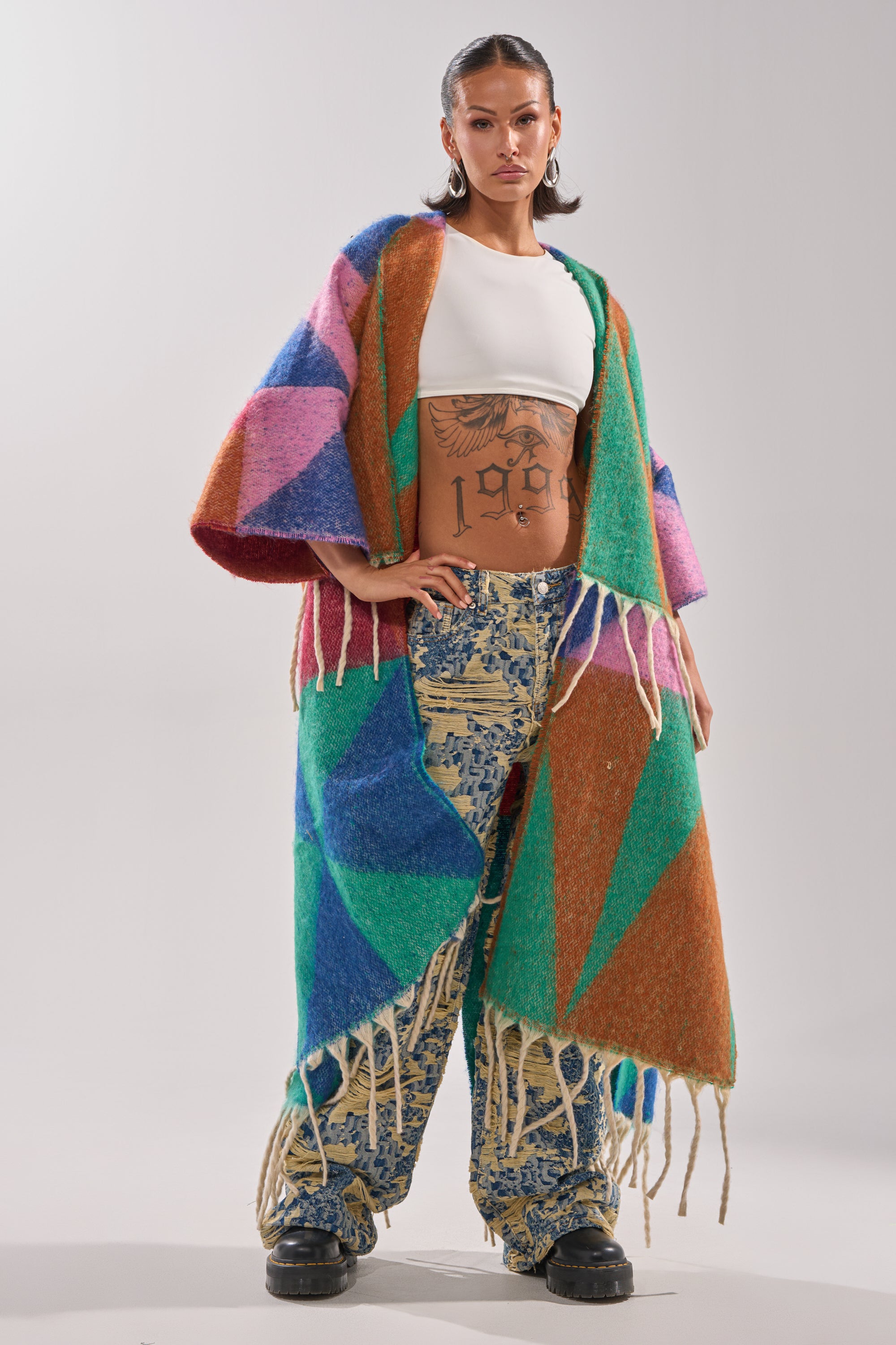 A woman exudes confidence in playful pigtails, large hoop earrings, tattoos on her midriff, a white crop top, and the RENEWED DREAM TAPESTRY PANT—featuring a two-tone wide-leg silhouette with colorful geometric patterns and fringed detail.