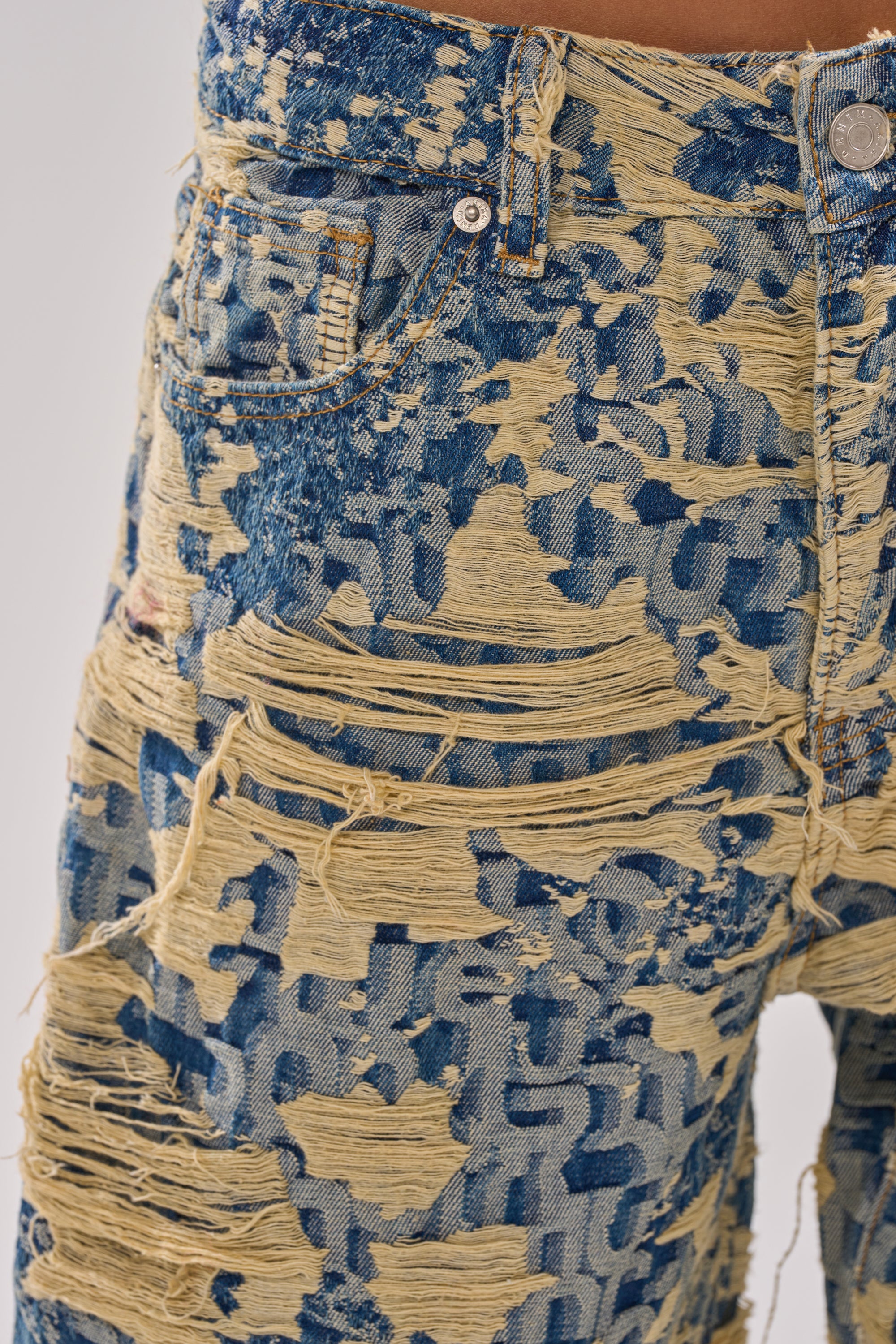 Close-up of RENEWED DREAM TAPESTRY PANT in blue and beige two-tone denim, showcasing heavy fraying, rips, exposed threads, a bold shredded pattern, and a metal waist button.