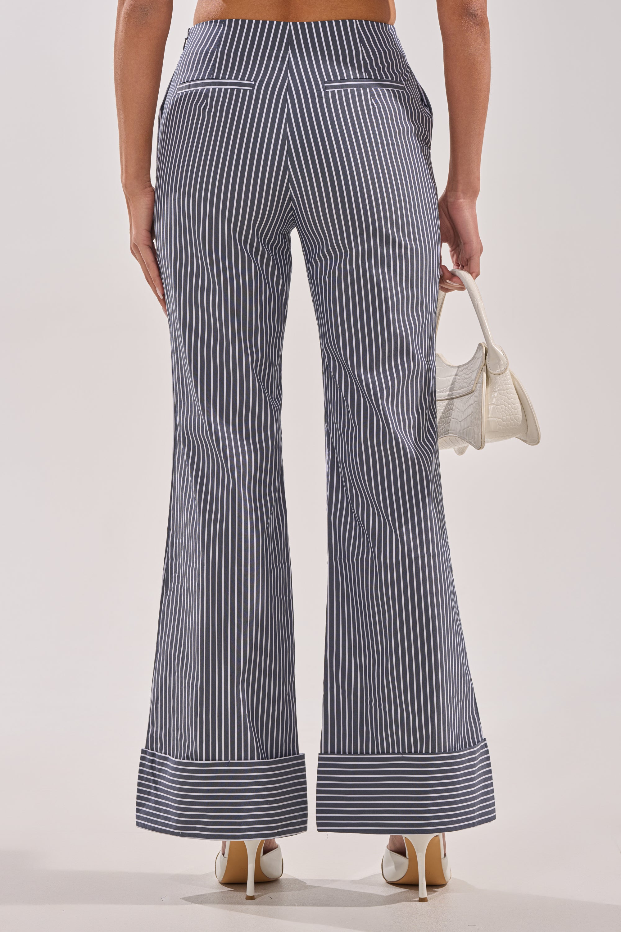A person is seen from behind wearing the MISS POPULAR POPLIN TROUSER—navy, high-rise tailored pants with wide white cuffs and vertical stripes—paired with a small white handbag and white high-heeled shoes. 4