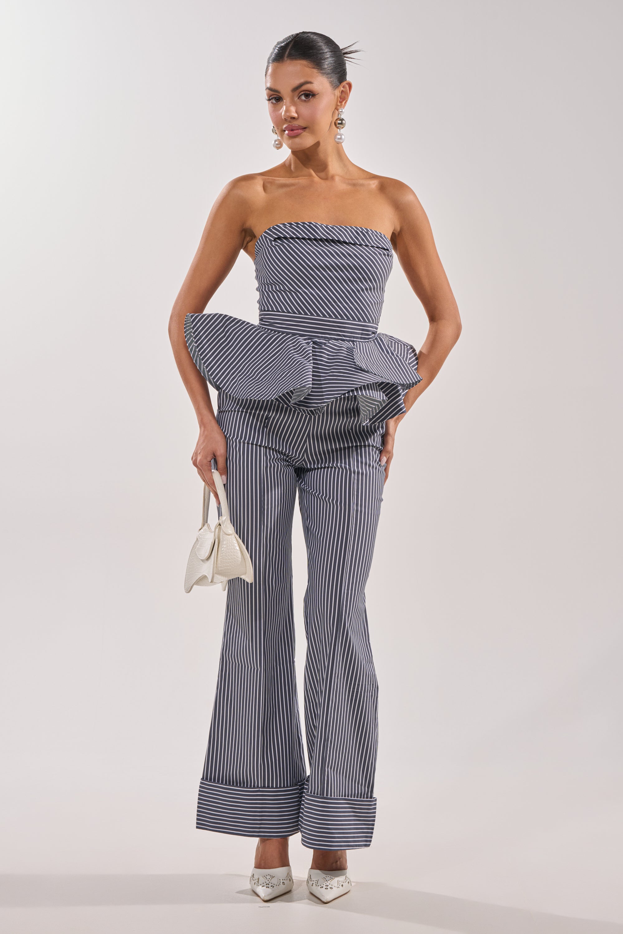 A woman poses holding a white handbag, wearing the MISS POPULAR POPLIN TROUSER—a strapless, navy and white pinstripe jumpsuit with peplum detail and high-rise waistband—paired with white pointed heels and statement earrings. 5