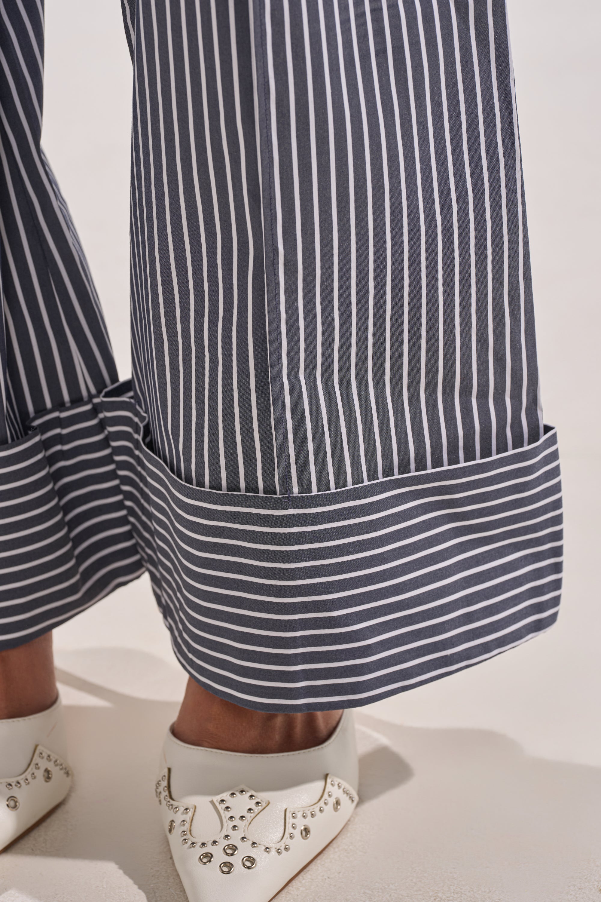 Close-up of a person wearing the MISS POPULAR POPLIN TROUSER, navy tailored wide-leg pinstripe pants with thick horizontal-striped cuffs, paired with white pointed-toe shoes adorned with silver studs. 6