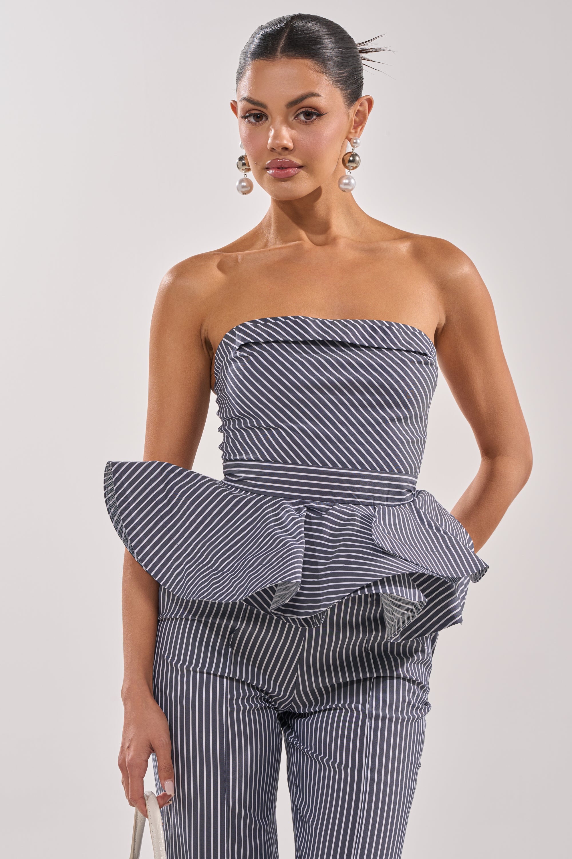 A woman wears the MISS POPULAR POPLIN CORSET in navy pinstripes with matching pants, styled with slicked-back hair, pearl drop earrings, and a small bag against a plain light background. 1