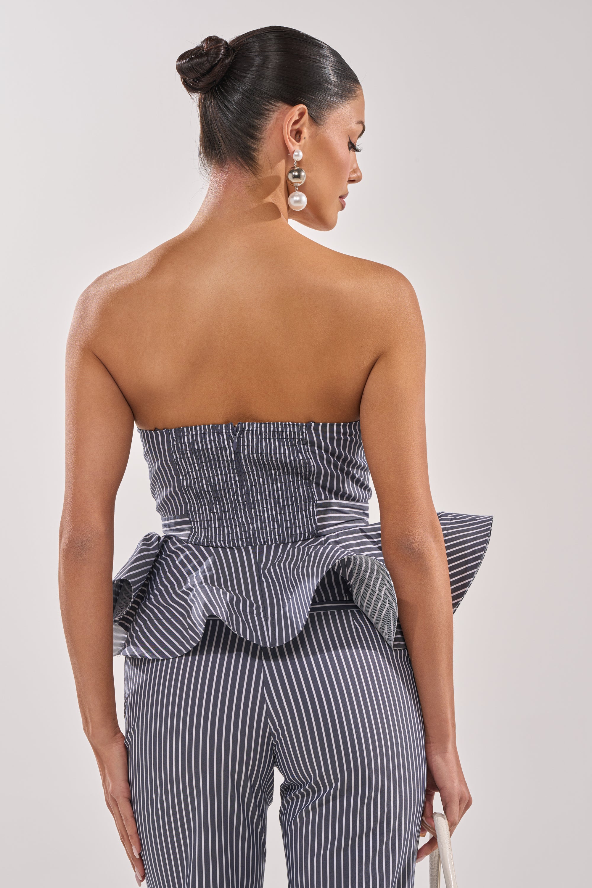 A woman with her hair in a bun models the MISS POPULAR POPLIN CORSET—a strapless, black-and-white pinstriped peplum style—with matching pants. Facing away, she carries a small white bag and wears bold pearl earrings for a chic look. 3