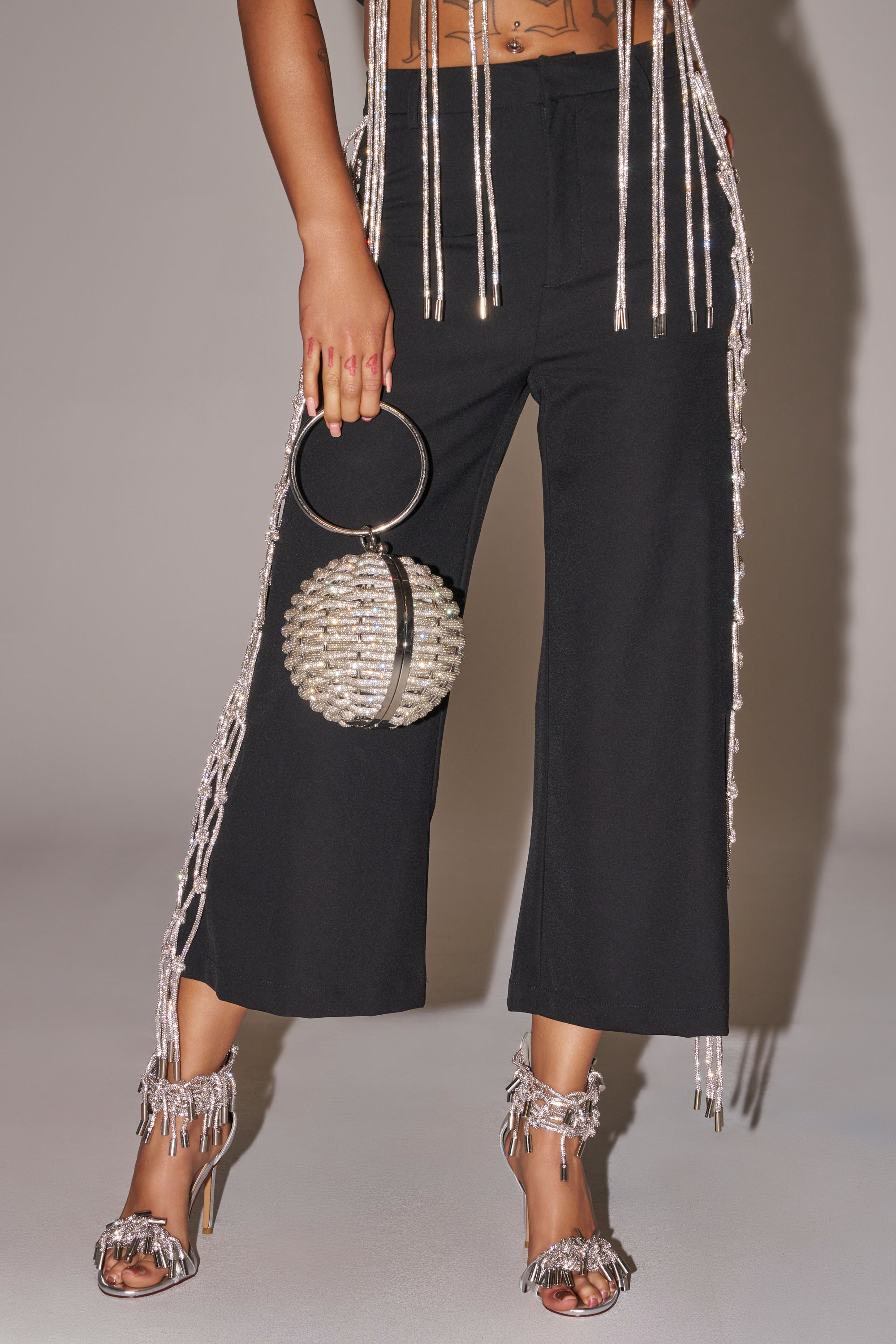 Wearing NINA RHINESTONE CULOTTES and metallic fringe high heels, a person holds a round beaded handbag with a large circular handle, while strands of metallic fringe drape from their top and shoes. 1