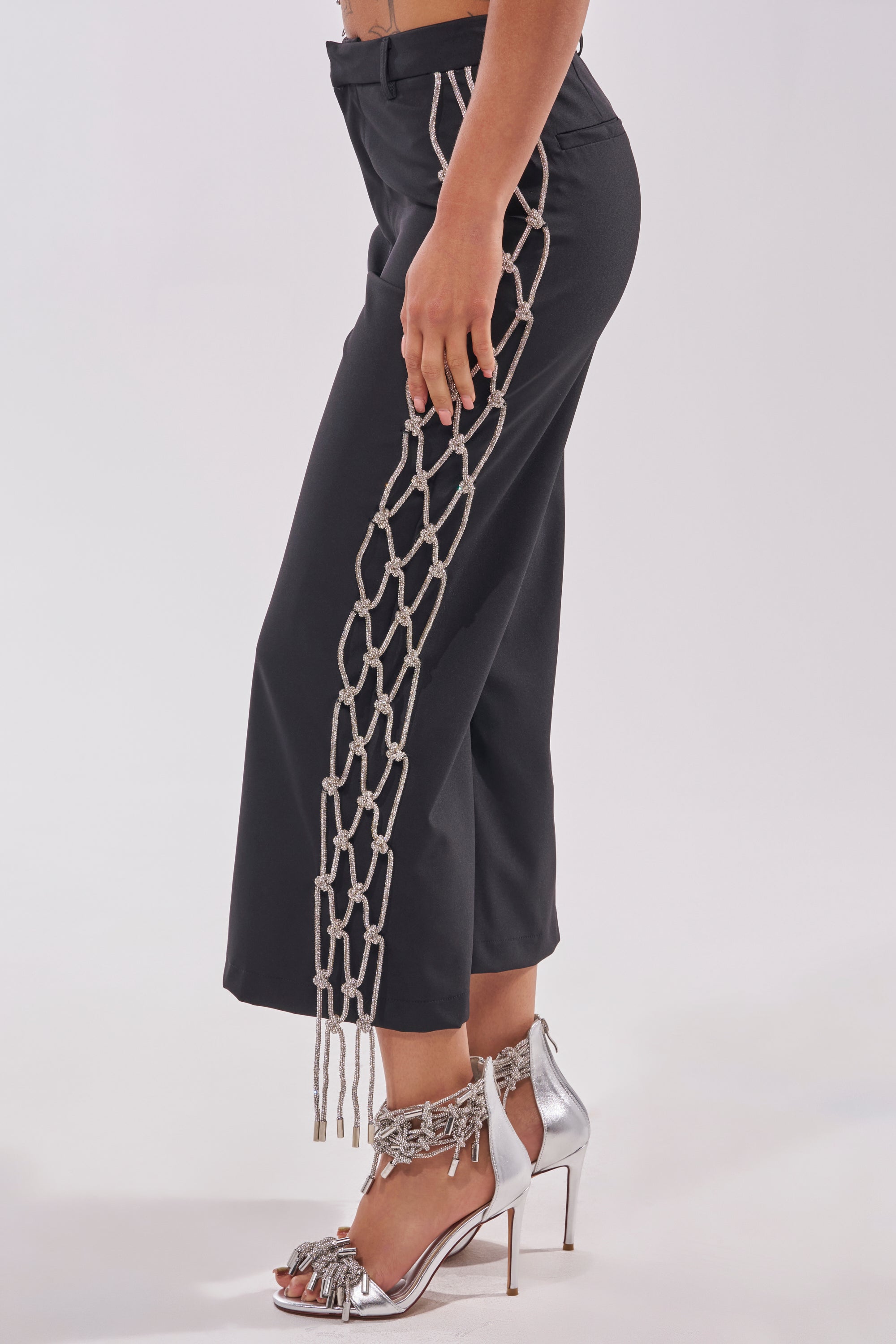 A person stands against a light background wearing stylish NINA RHINESTONE CULOTTES with knotted rope detailing along the side seam and silver tasseled high-heeled sandals. 3
