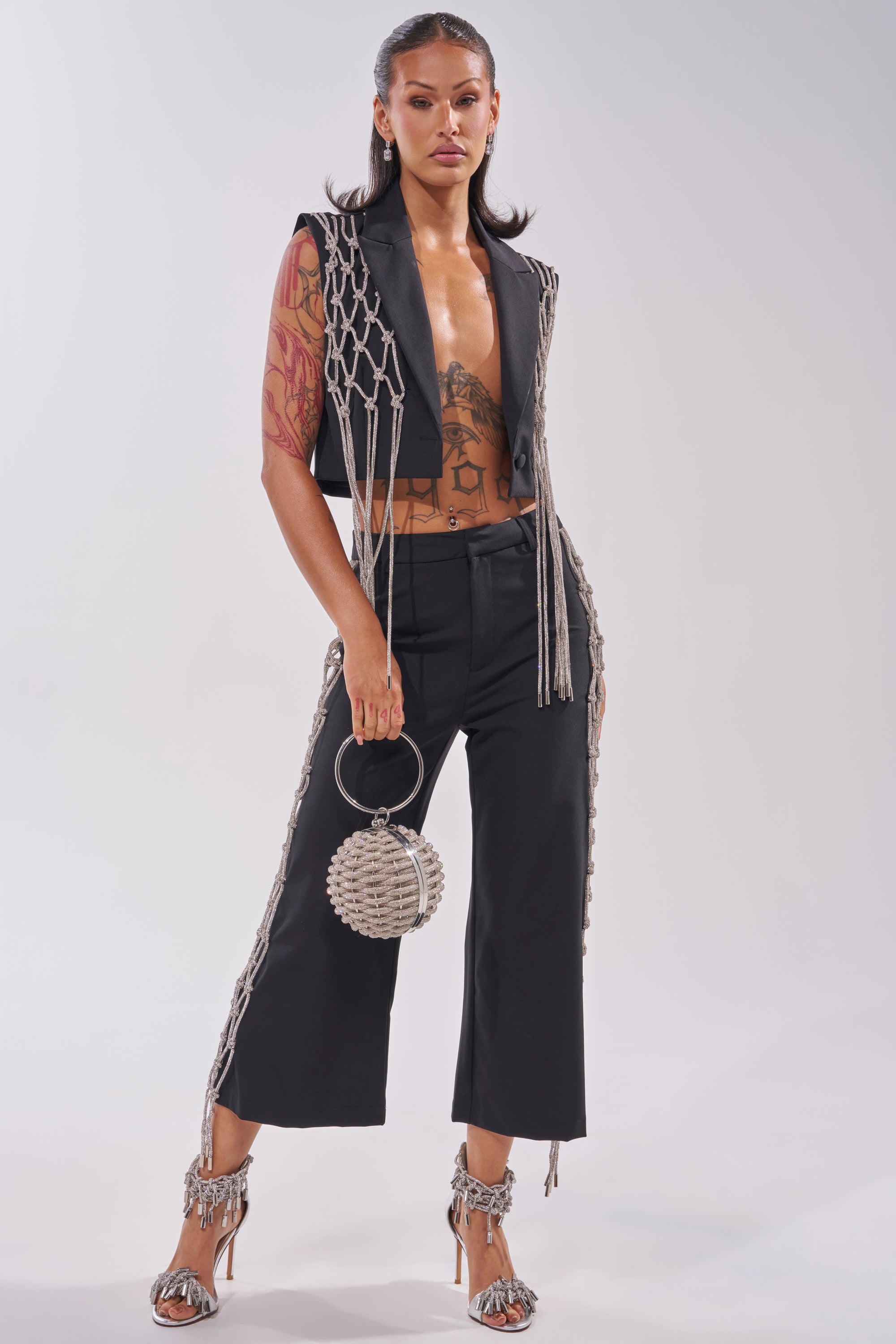 A woman exudes confidence in the NINA RHINESTONE CULOTTES, paired with a netted vest and round beaded handbag. She completes her look with open-toed tassel heels, slicked-back hair, and visible tattoos on her arms and torso. 5