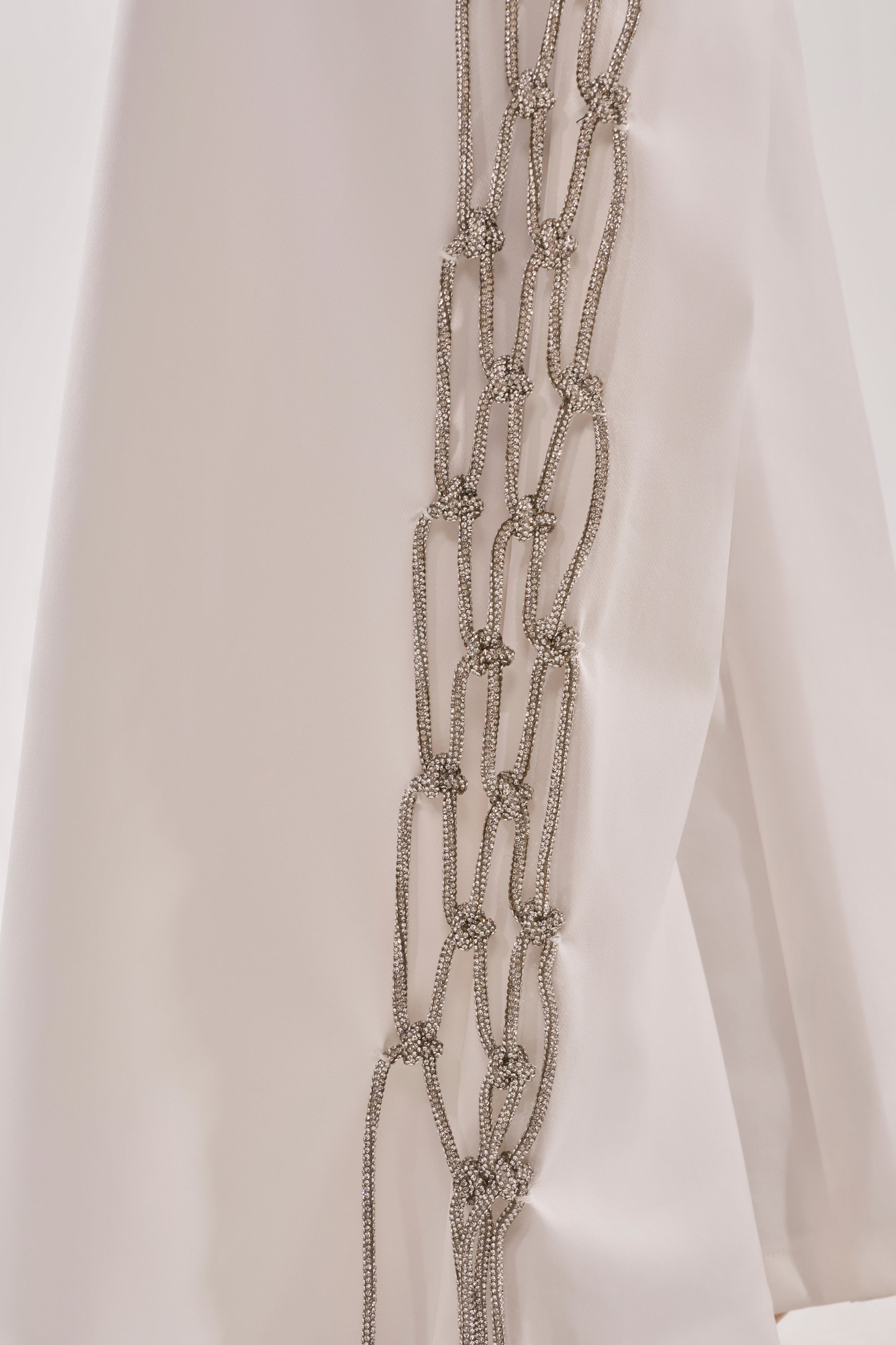 Close-up of the BELLISSIMA EMBELLISHED TROUSER, showcasing silver chain details intricately knotted and arranged vertically on white fabric for a decorative, standout look. 9