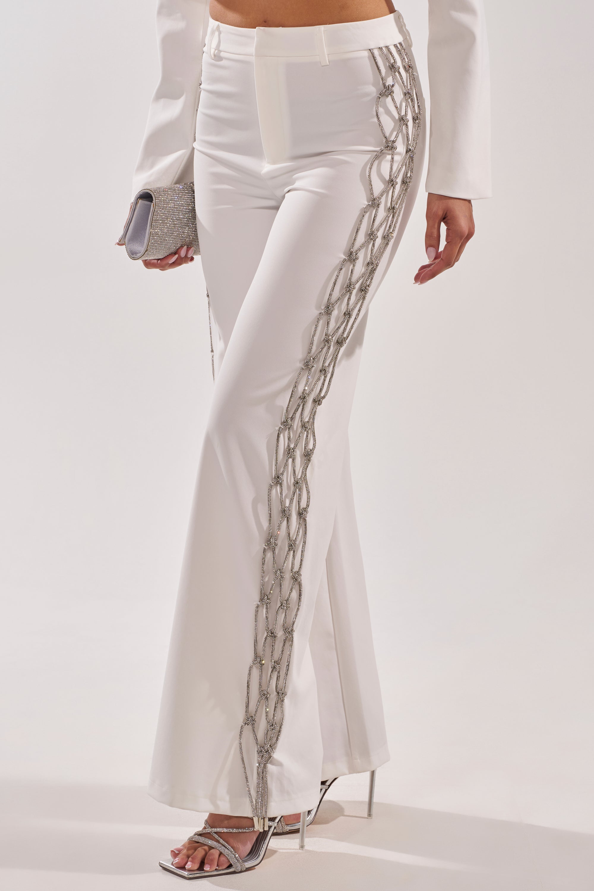 A person wearing the BELLISSIMA EMBELLISHED TROUSER, featuring silver chain details along the sides, paired with a small silver clutch and high-heeled sandals. Image is cropped from the waist down. 1