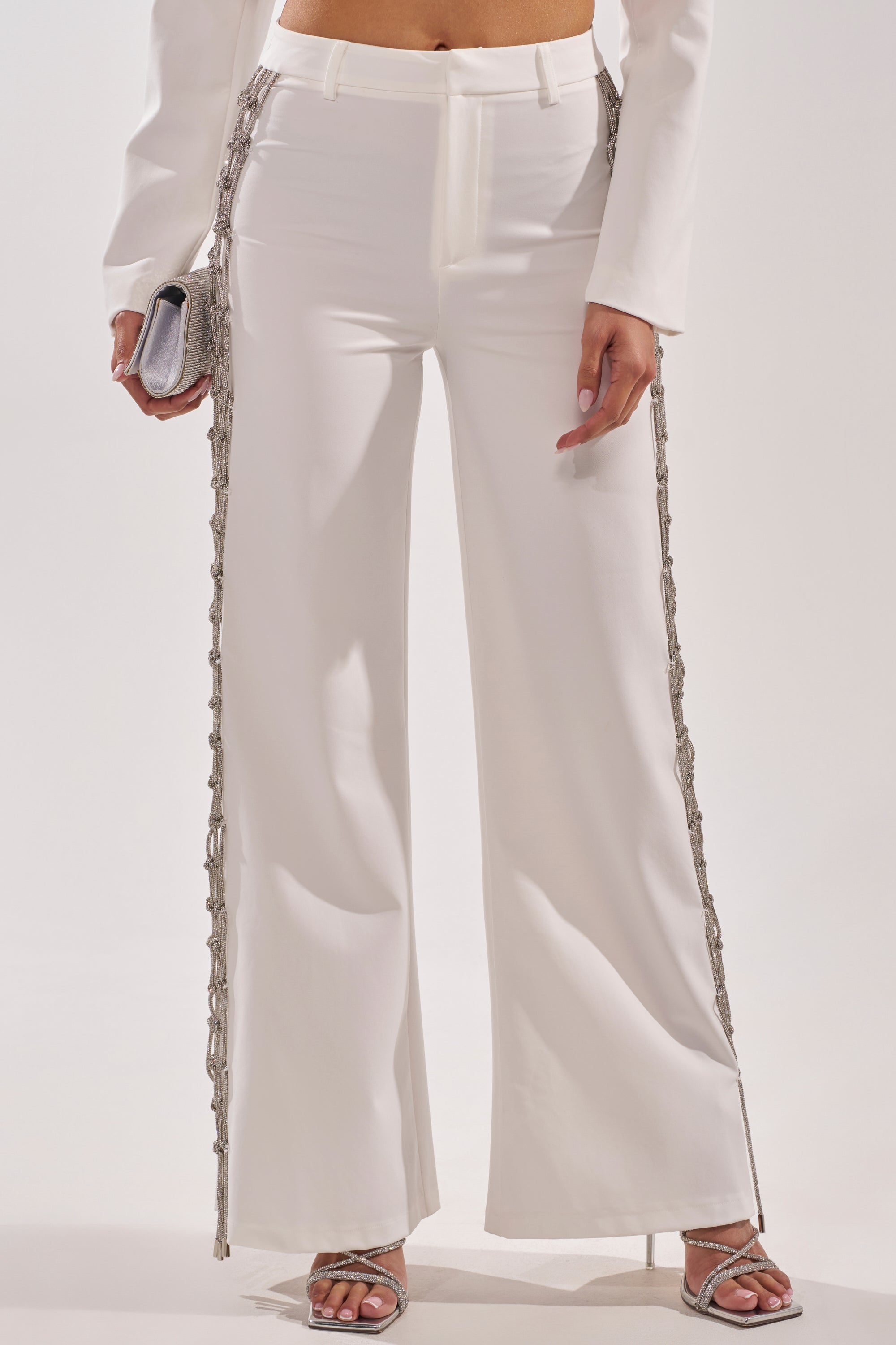 A person wearing BELLISSIMA EMBELLISHED TROUSER—high-waisted white pants with silver chain details—paired with a matching top, silver high-heeled sandals, and a small silver clutch. 3