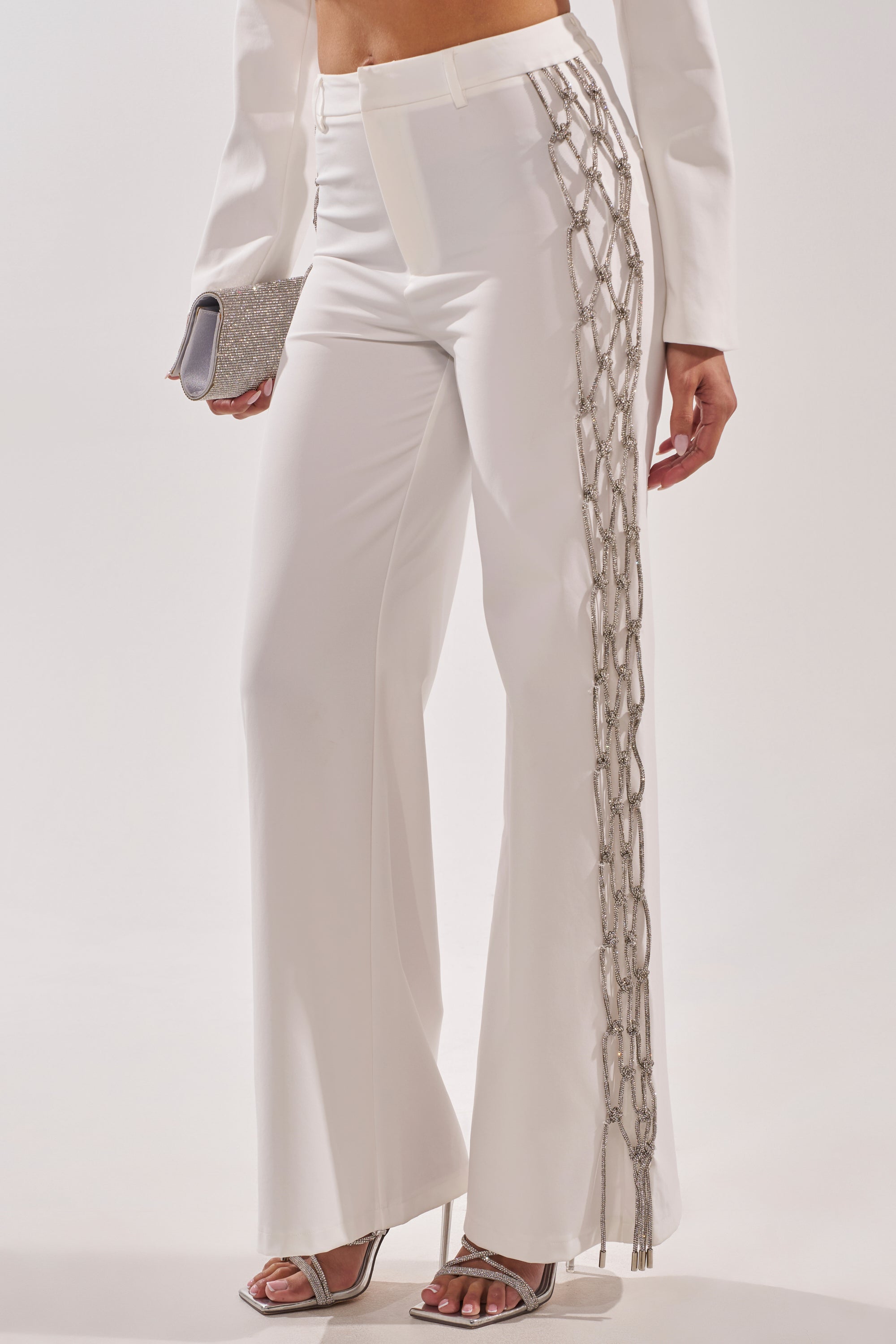A person wearing the BELLISSIMA EMBELLISHED TROUSER—white high-waisted pants with crystal details and silver chain accents—paired with a white blazer, silver heels, and a matching silver clutch. 4