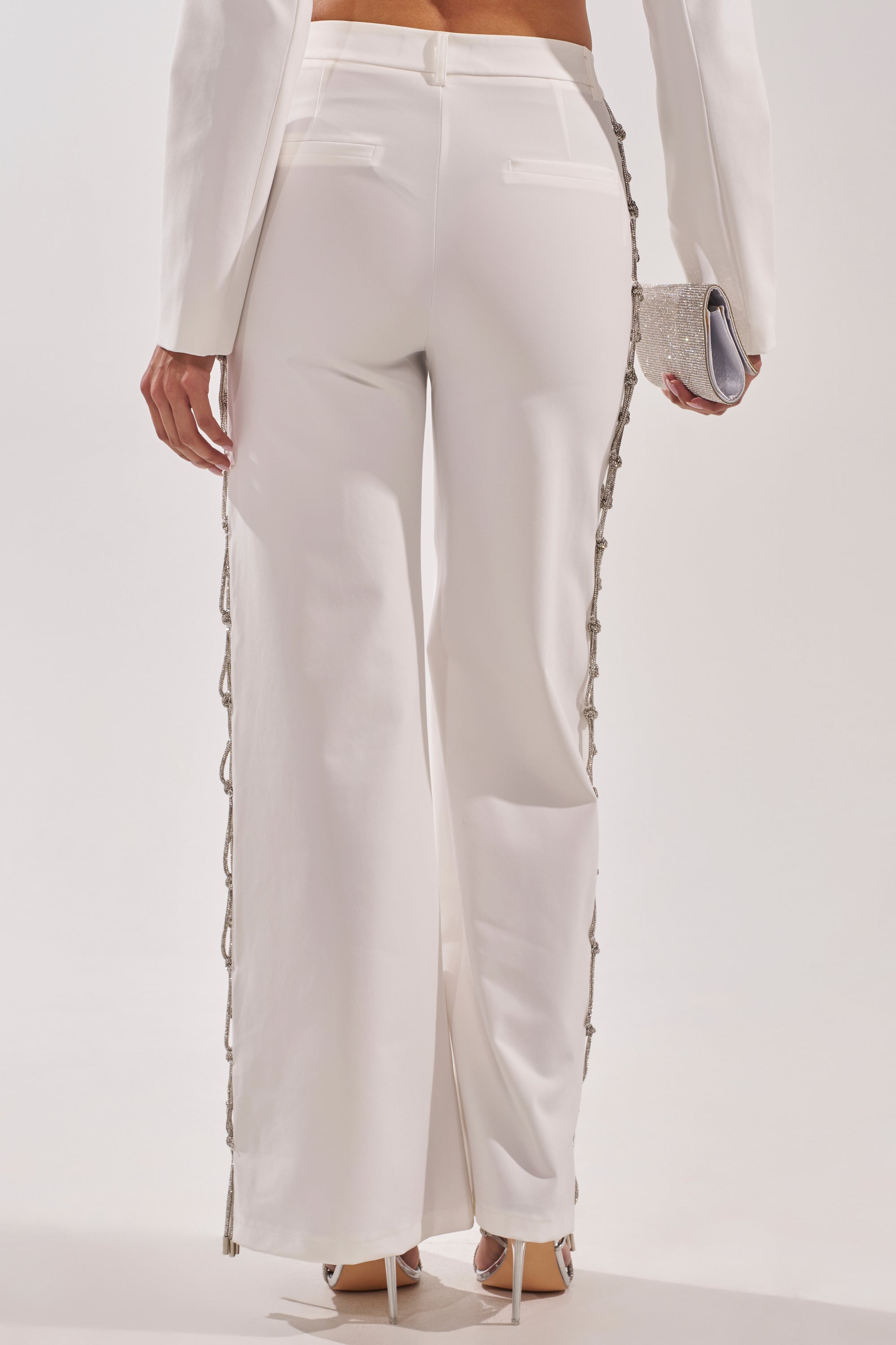A person is shown from the back wearing BELLISSIMA EMBELLISHED TROUSER—crystal embellished white pants with decorative side trim—paired with white heels and a small silver clutch against a plain background. 5