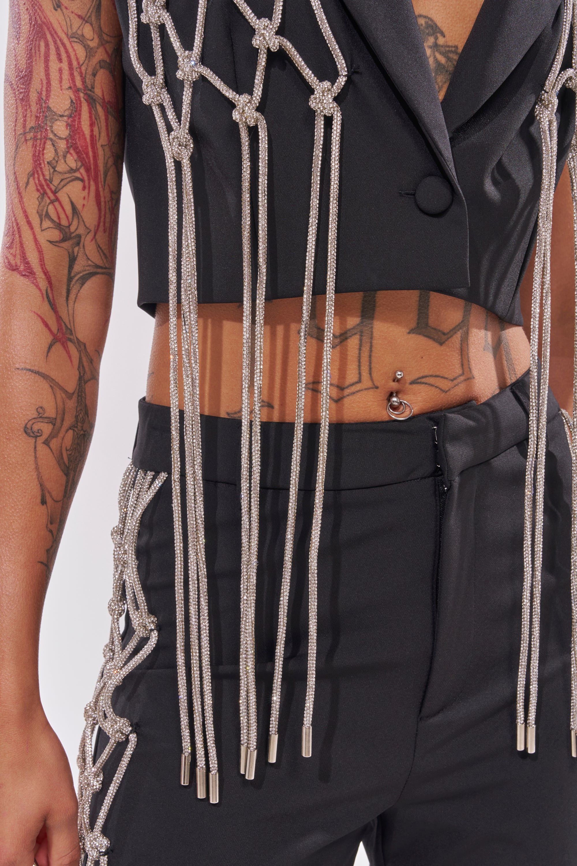 Wearing the NINA RHINESTONE BLAZER VEST and coordinating pants with silver rope accents, this look shows off your midriff, belly piercing, and tattoos—ideal for making a bold statement on a night out. 10