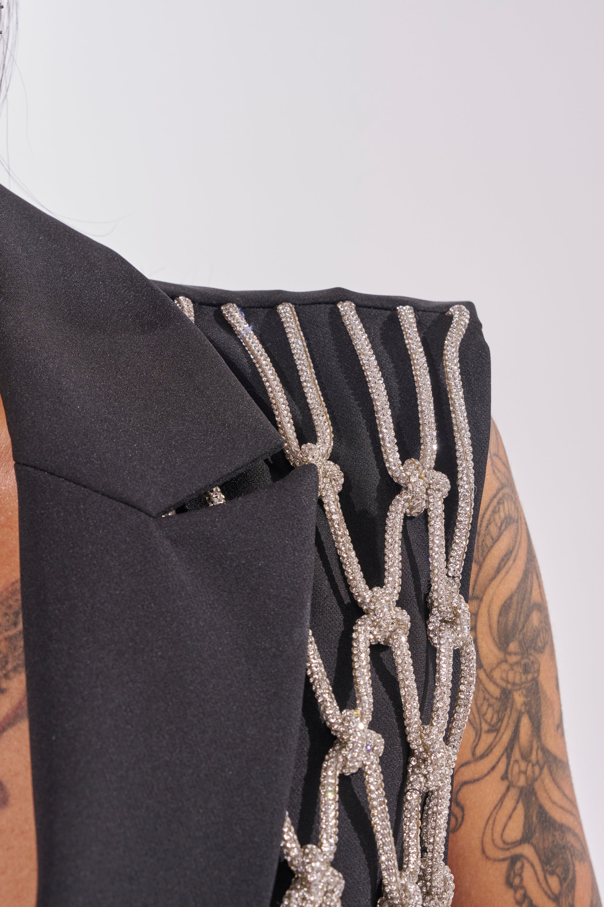 Close-up of a person wearing the NINA RHINESTONE BLAZER VEST, featuring dazzling silver rope details on black fabric. Part of their shoulder, tattooed arm, and the vest’s structured collar are visible—ideal for a bold night out look. 11