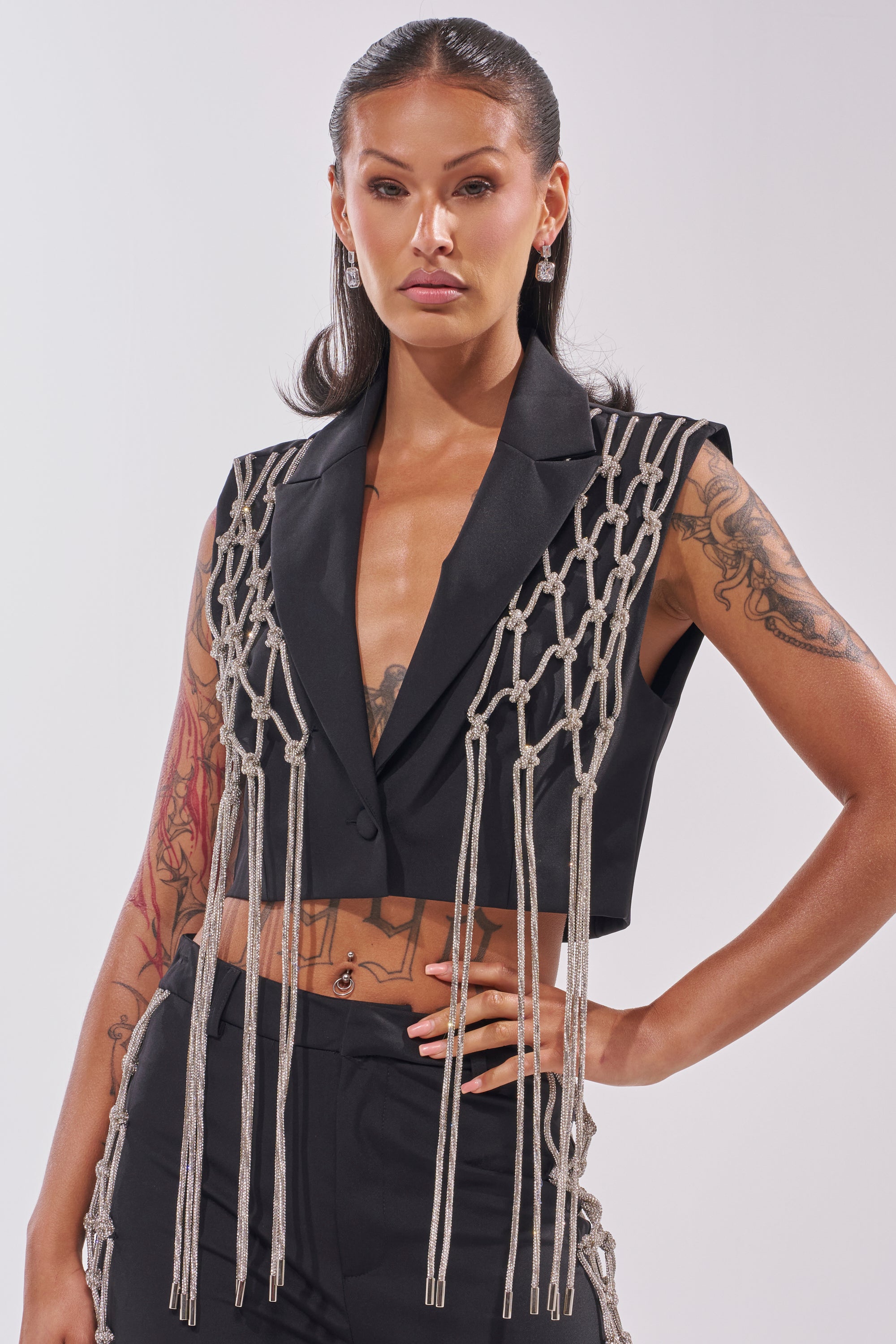 A woman with long dark hair and tattoos models the NINA RHINESTONE BLAZER VEST, a sleeveless black cropped blazer featuring knotted silver chains, posing confidently against a light background—perfect for making a statement. 1
