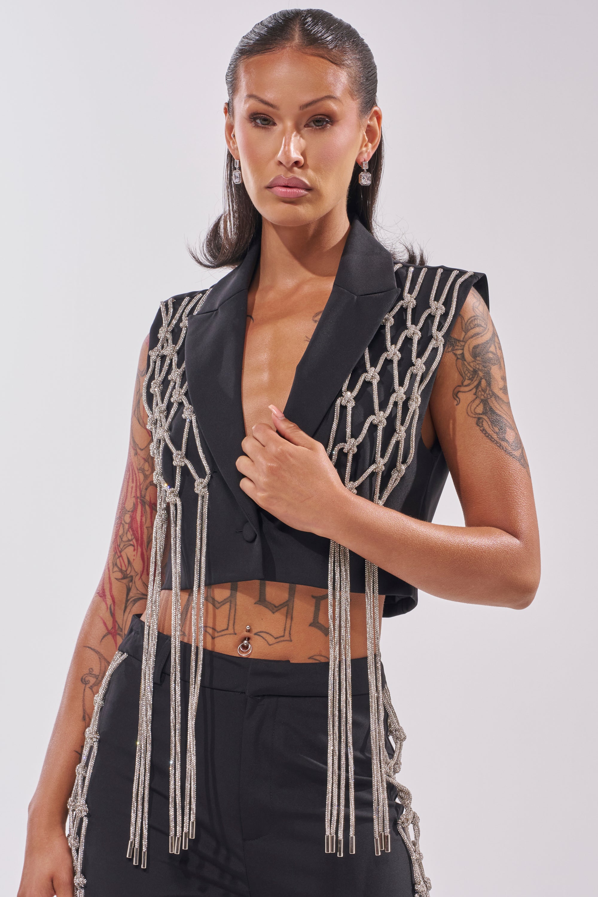 A woman with long dark hair, tattoos, and a navel piercing models the NINA RHINESTONE BLAZER VEST—a bold black sleeveless blazer with striking rhinestone rope details—against a plain light background. 5