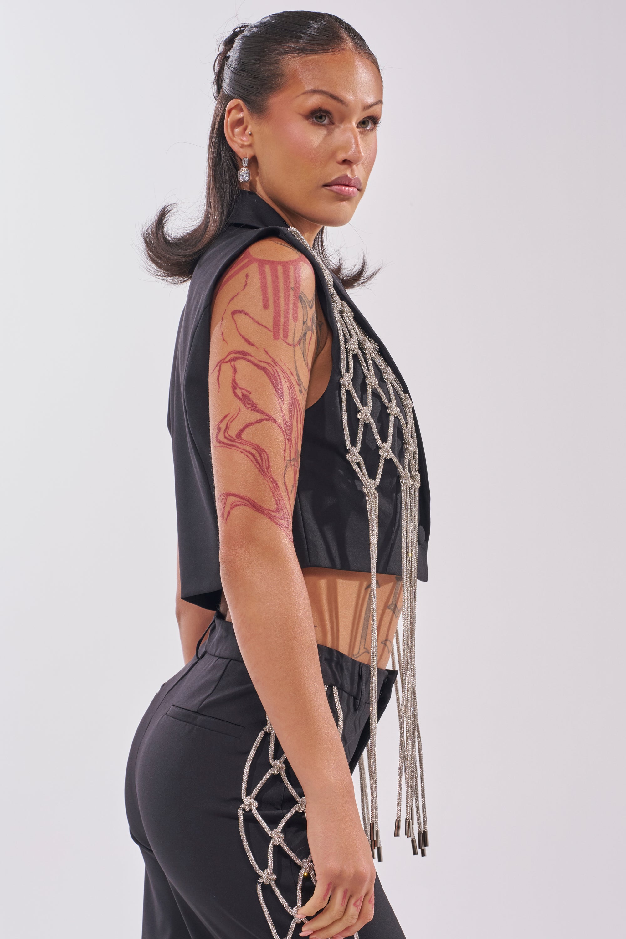 A confident woman with long dark hair models the NINA RHINESTONE BLAZER VEST and black pants featuring rope details, showcasing her bold red abstract arm tattoo against a plain background—perfect for a statement night out. 6