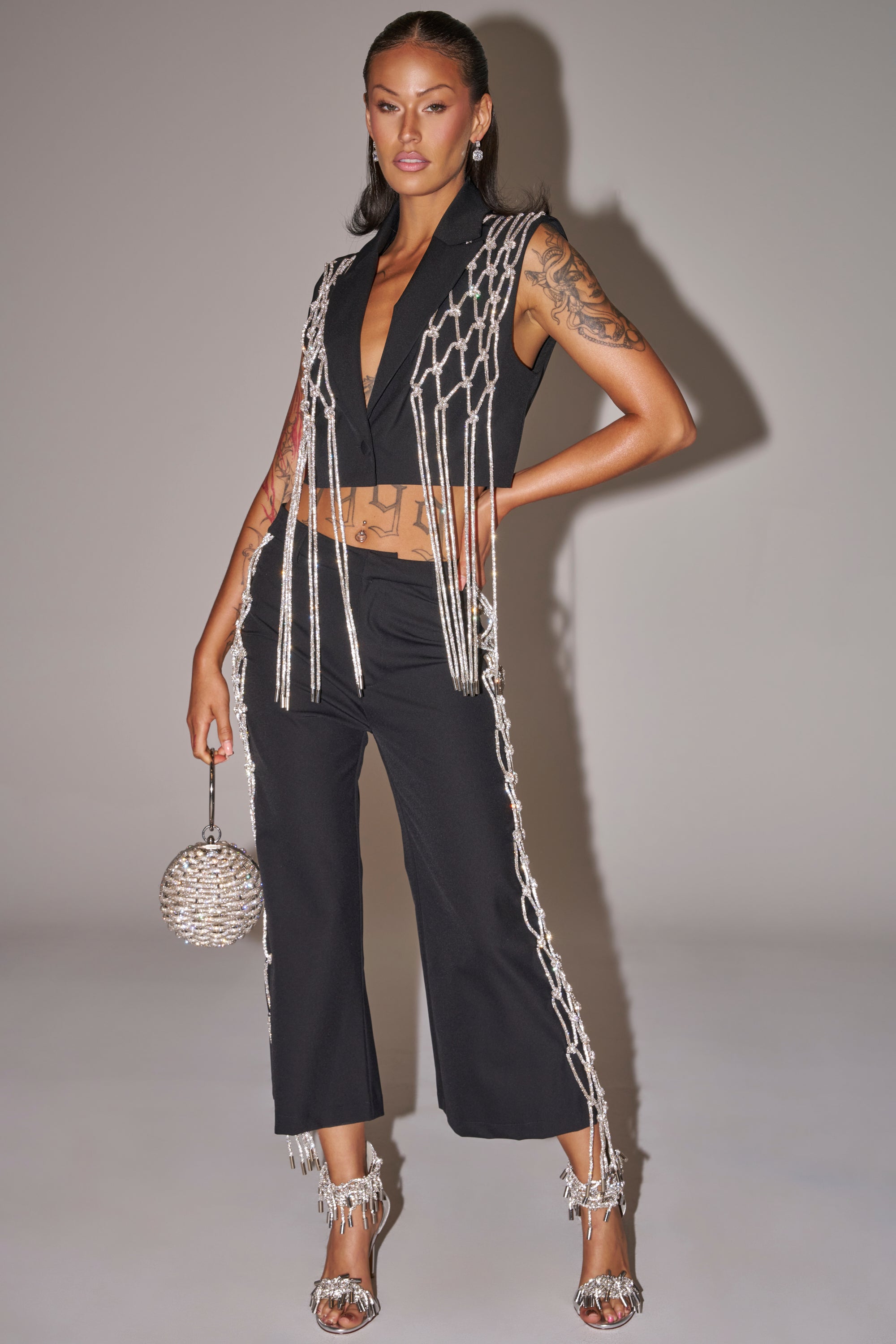 A woman wears the NINA RHINESTONE BLAZER VEST with cropped black pants featuring silver chain fringe, pairing the statement look with a round silver handbag, silver-fringe heels, and showcasing her arm tattoos for a bold night-out style. 8