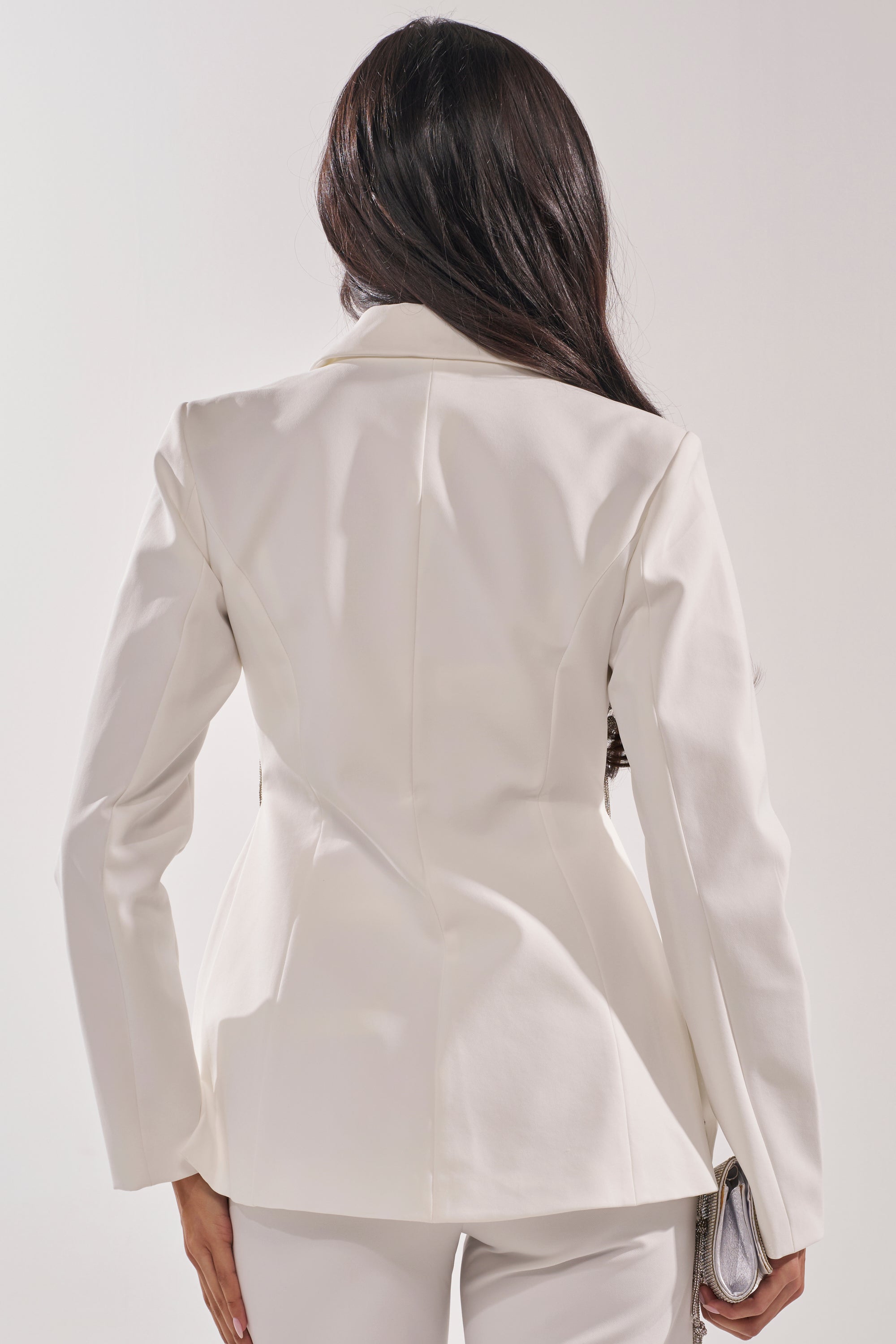 A woman with long dark hair is seen from behind wearing the BELLISSIMA EMBELLISHED BLAZER and matching pants, holding a small clutch in her left hand against a plain background. 4