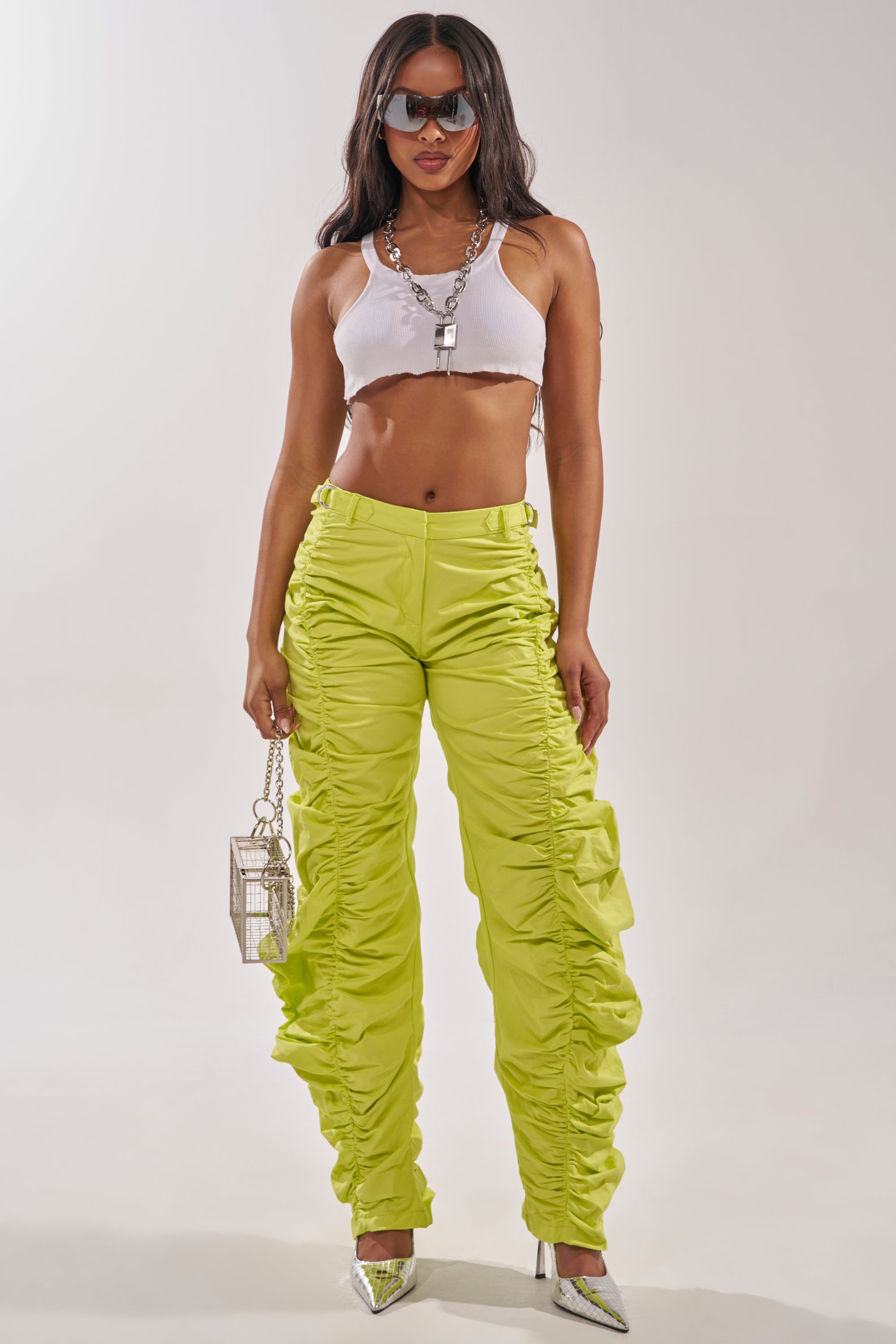 A woman stands confidently in a white crop top, silver heels, sunglasses, and the NOT IN THE MOOD RUCHED TROUSER—bold, high-waisted statement pants. She accessorizes with a small silver geometric handbag and layered necklaces. 0