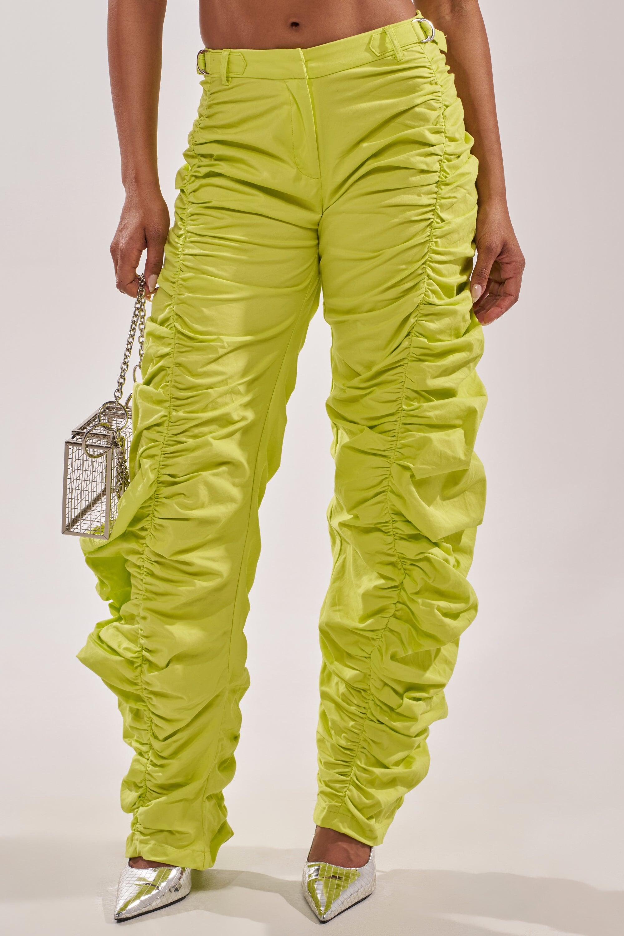 Wearing the NOT IN THE MOOD RUCHED TROUSER in green with ruffled side detailing, this look is paired with a small metallic purse and silver pointed-toe heels. These bold festival pants are shown from the waist down. 1