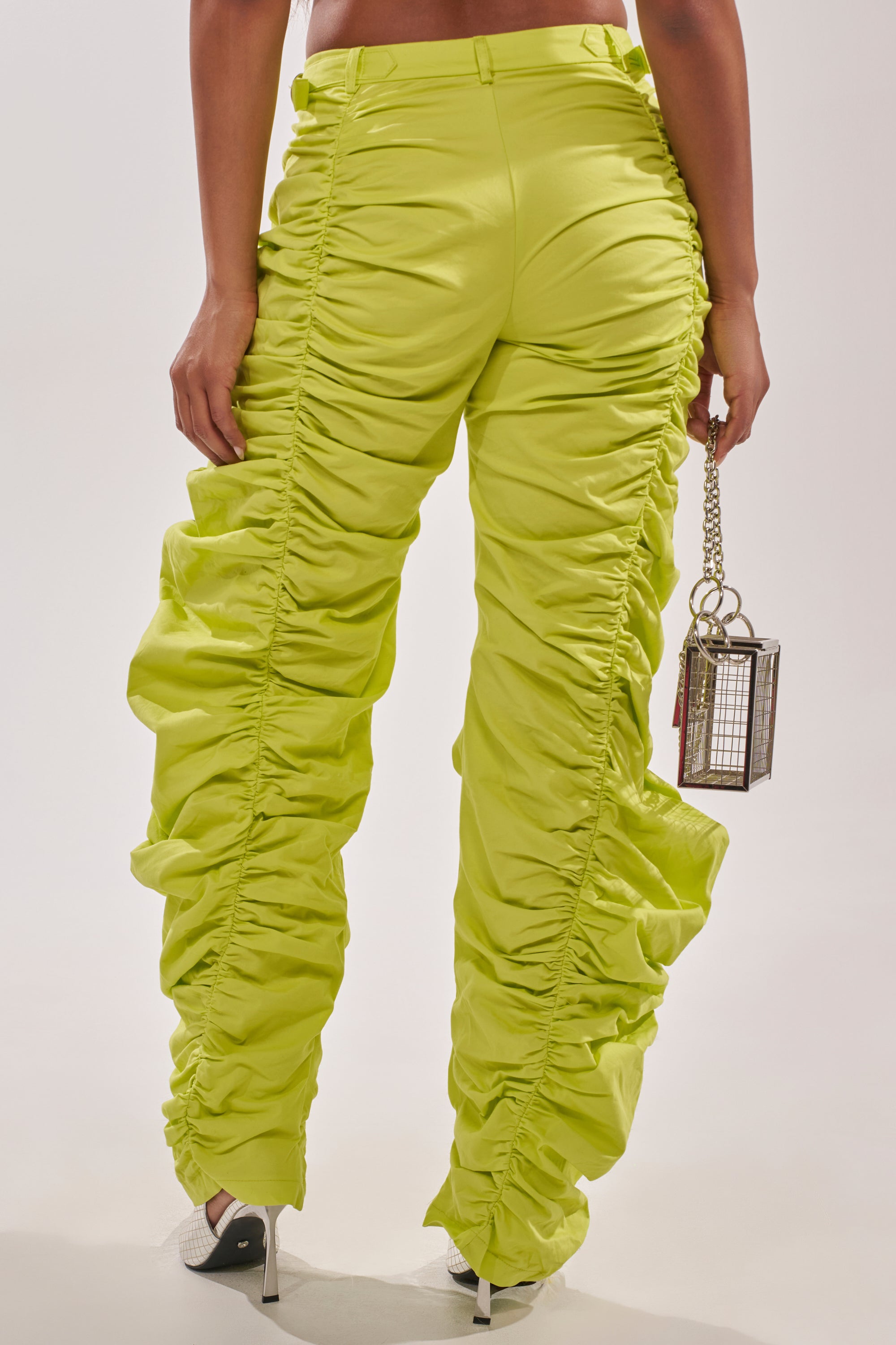 A person stands facing away, wearing the NOT IN THE MOOD RUCHED TROUSER in green with a high-rise waist and white heeled shoes, holding a small metallic box-shaped handbag against a plain light background. 3