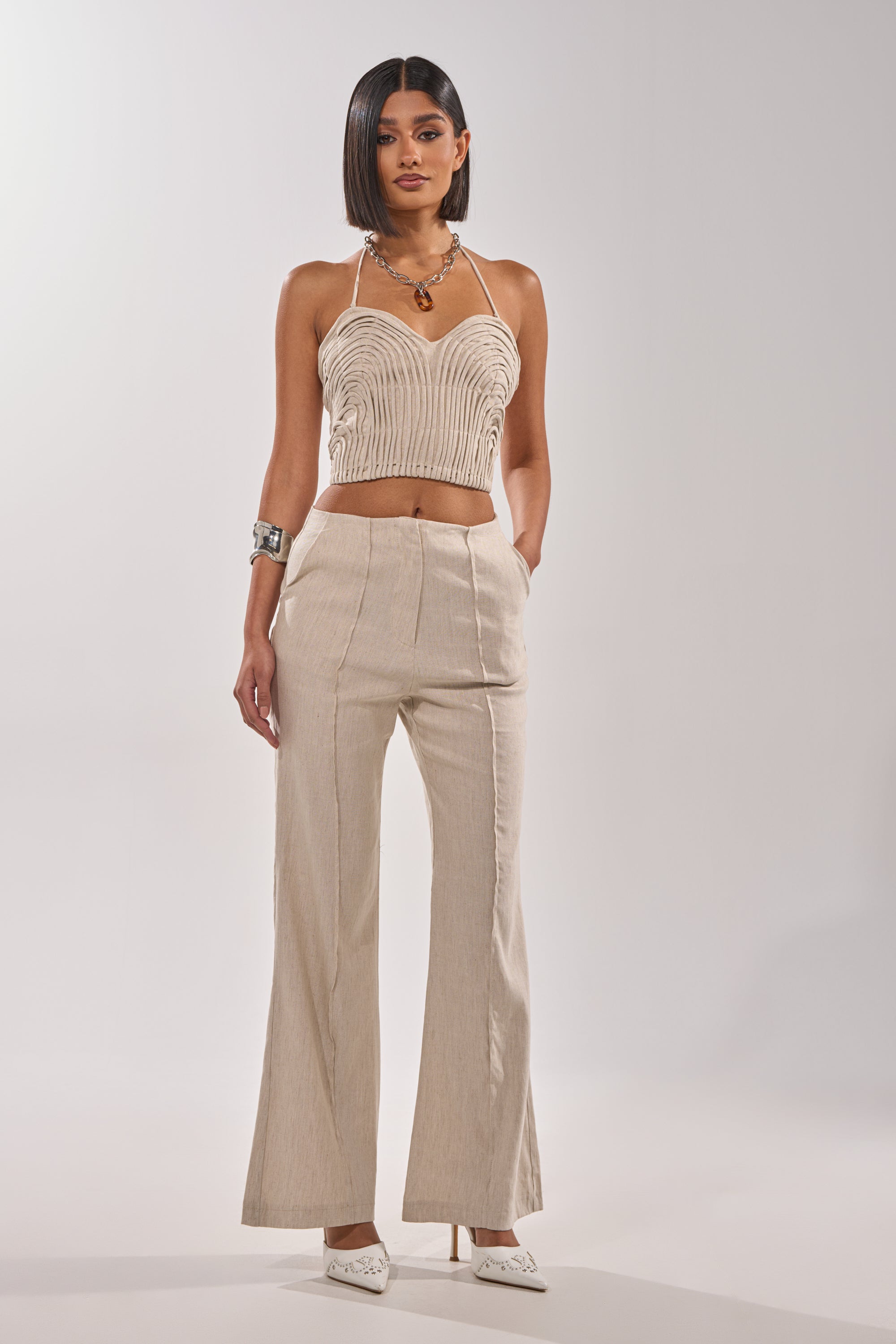 A woman stands against a plain background wearing the LAY IT LOW TROUSER, high-rise wide-leg linen pants in beige, paired with a matching sleeveless halter crop top. She completes the minimalist look with a necklace, bracelet, and white heels. 0