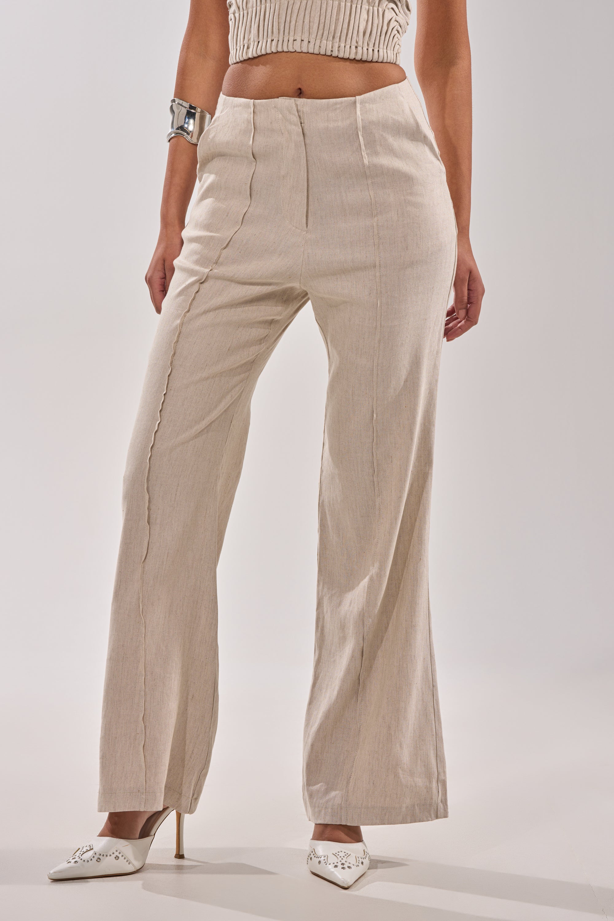 Woman styled in LAY IT LOW TROUSER—high-rise, wide-leg linen pants with vertical seams—paired with a cream knit crop top, silver bracelet, and white pointed-toe heels for a minimalist look against a plain light background. 1