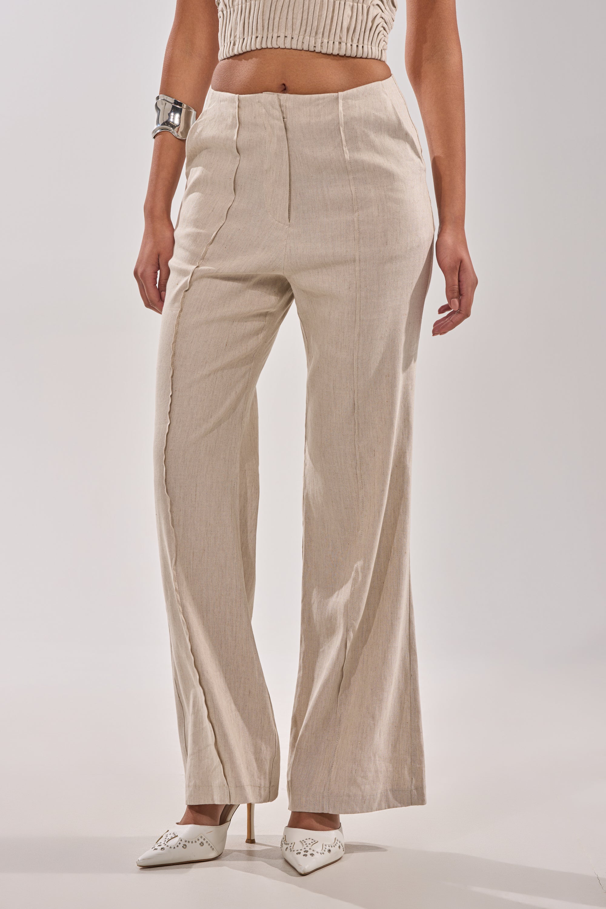 Wearing the LAY IT LOW TROUSER, a woman in wide-leg linen pants with a high-rise waist pairs them with a ribbed crop top, white pointed-toe heels, and a chunky silver bracelet, showcasing refined minimalist style against a plain light background. 2