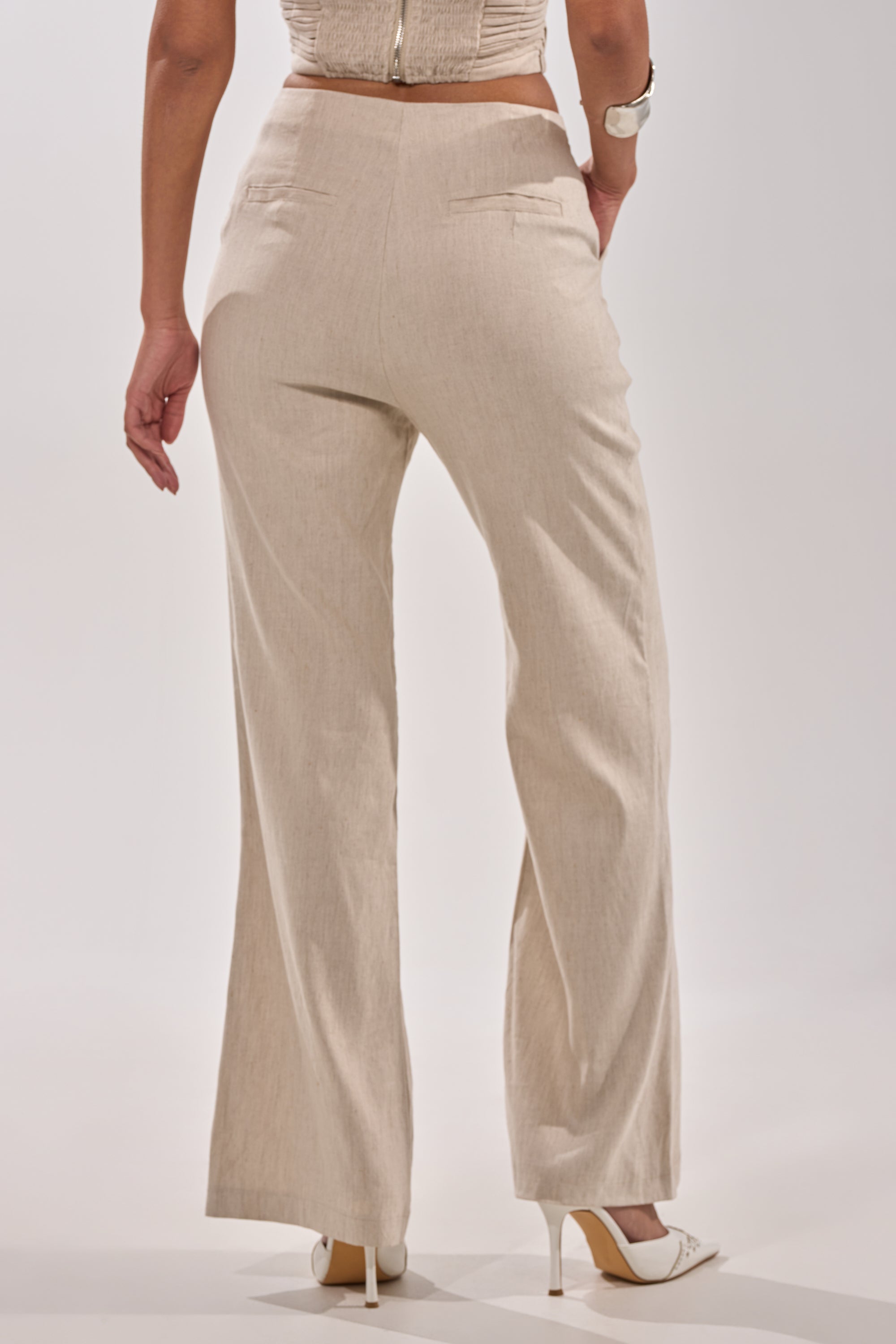 A woman, shown from behind against a plain white backdrop, models the LAY IT LOW TROUSER—high-rise, wide-leg linen pants—paired with a cropped top and white high-heeled shoes for a minimalist look. 4