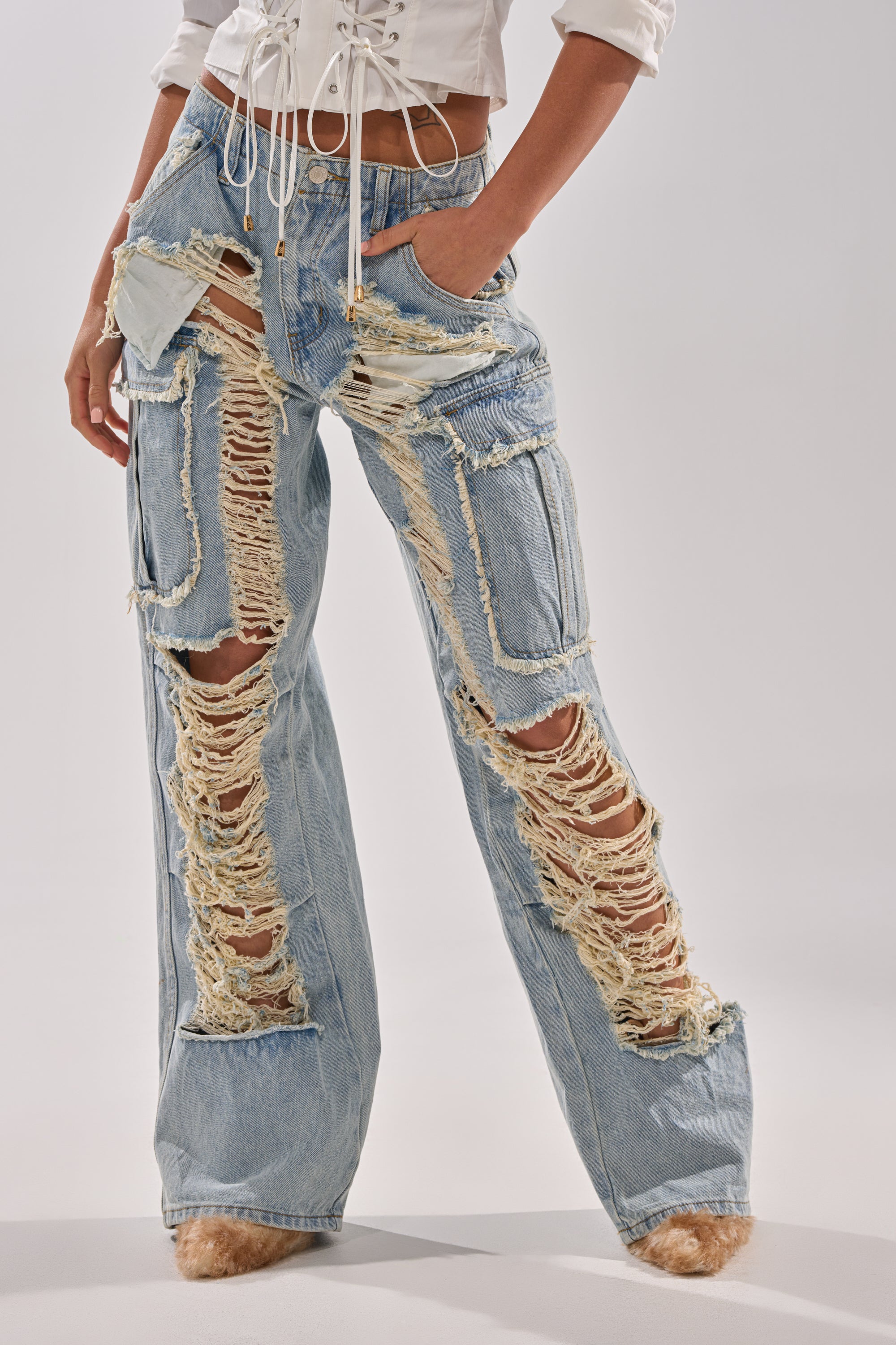 Wearing the ALMA DISTRESSED DENIM CARGO PANTS—light-wash, high-waisted jeans with large frayed holes—paired with a white cropped top, hands in pockets and feet slightly turned out.