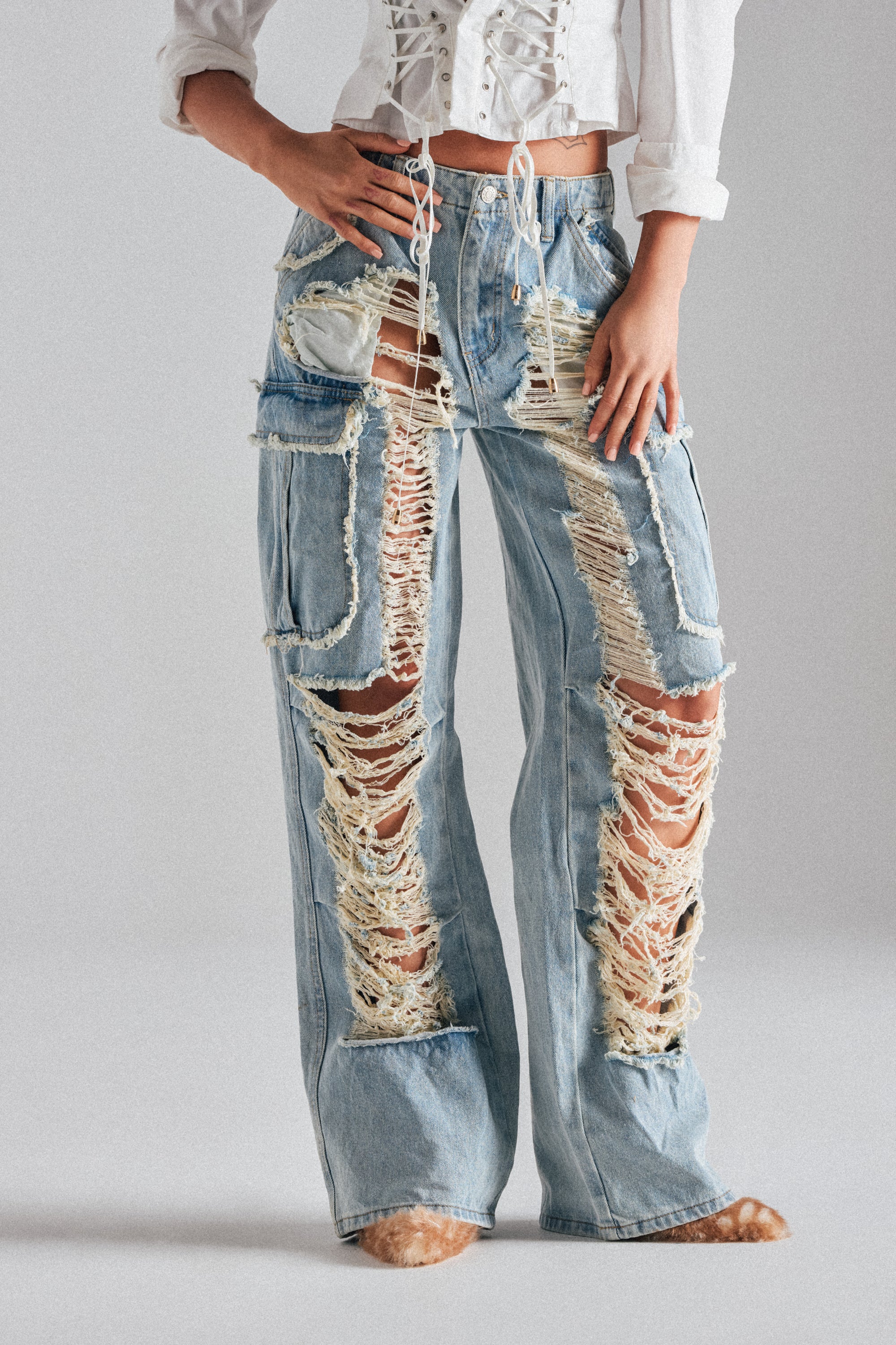 A person wears ALMA DISTRESSED DENIM CARGO PANTS featuring a light wash, large rips, and frayed edges with a white lace-up corset top. The high-waisted cargo pants and top combine for an edgy look; head and feet are out of frame.