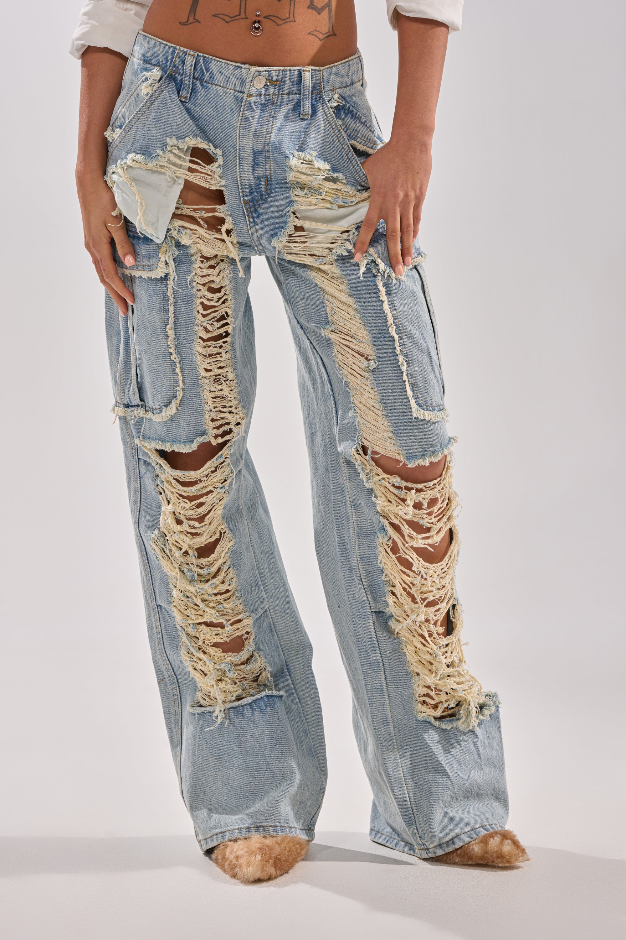 A person is shown from the waist down wearing ALMA DISTRESSED DENIM CARGO PANTS, featuring a light wash, large rips, and frayed holes on the thighs and knees, standing against a light background.