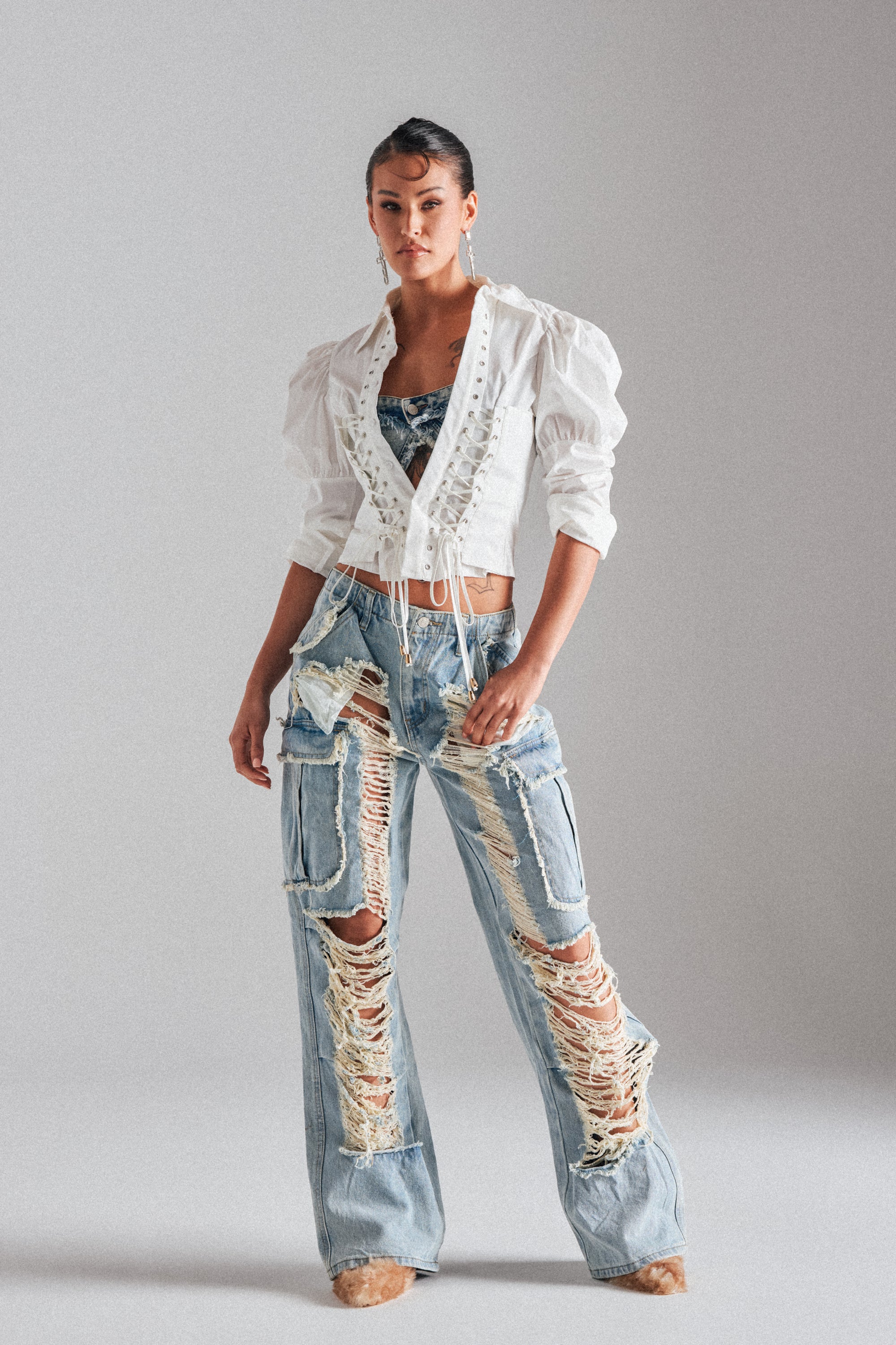 A woman stands against a plain backdrop wearing light-wash ALMA DISTRESSED DENIM CARGO PANTS with heavy rips and fraying, paired with a white, long-sleeved lace-up blouse and a patterned crop top.
