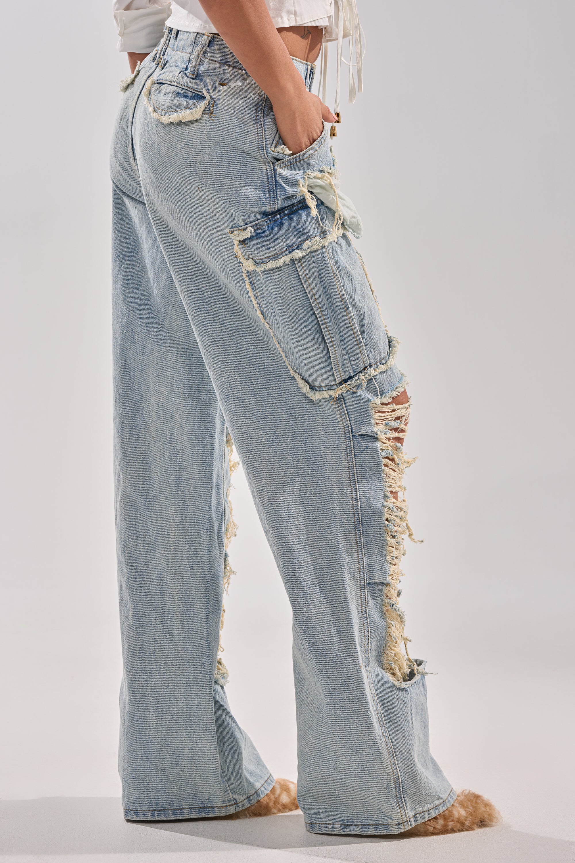 A person in ALMA DISTRESSED DENIM CARGO PANTS—light blue, high-waisted, featuring large side pockets and frayed distressing—stands with one hand in their pocket against a plain background.