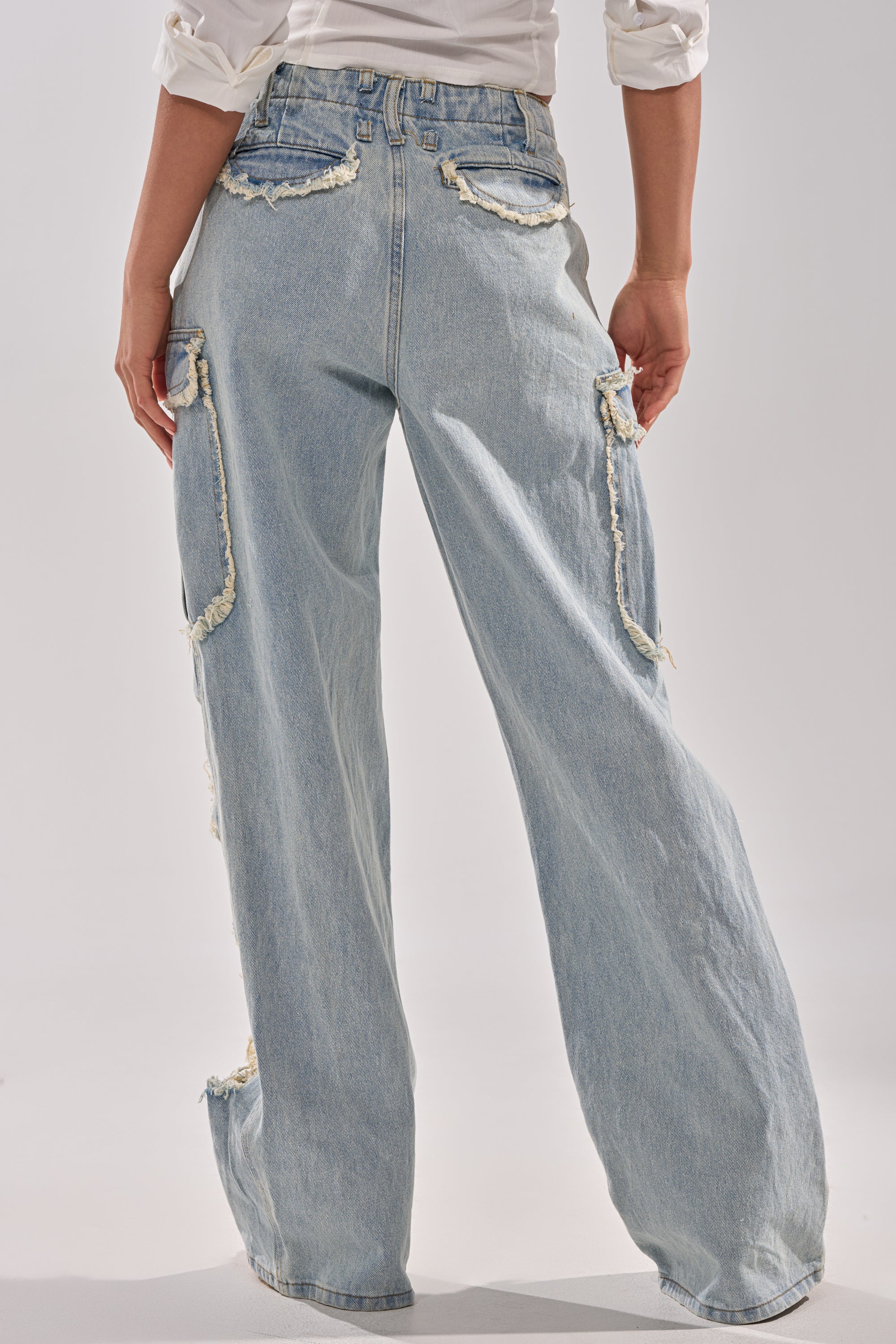 A person is seen from the back wearing ALMA DISTRESSED DENIM CARGO PANTS—light-wash, high-waisted, wide-leg jeans with frayed edges, multiple pockets, and a relaxed-fit style. The person's arms are bent at the elbows.