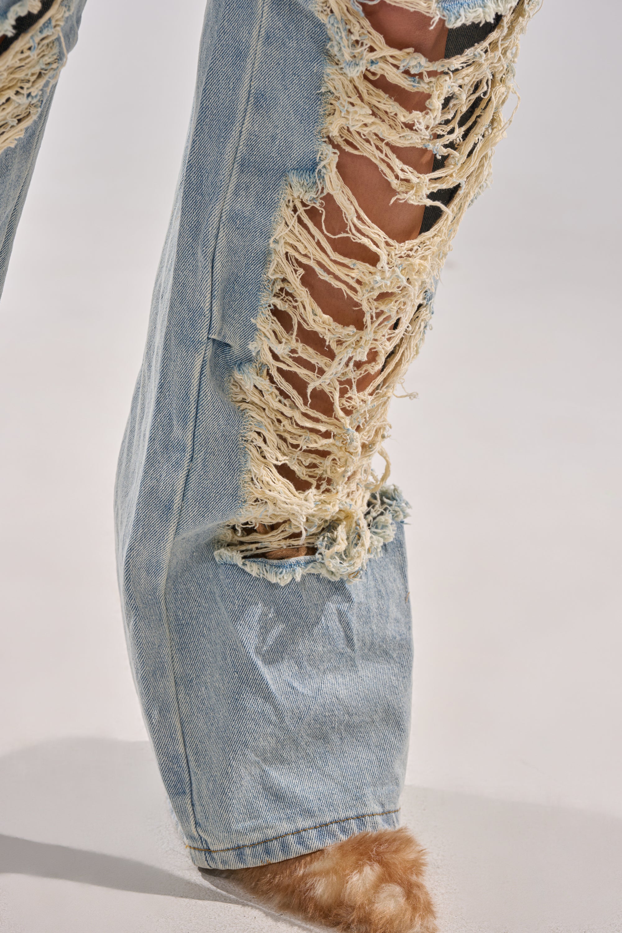A close-up of a person wearing ALMA DISTRESSED DENIM CARGO PANTS—high-waisted, light-wash jeans with large frayed holes—and fuzzy tan-colored footwear.