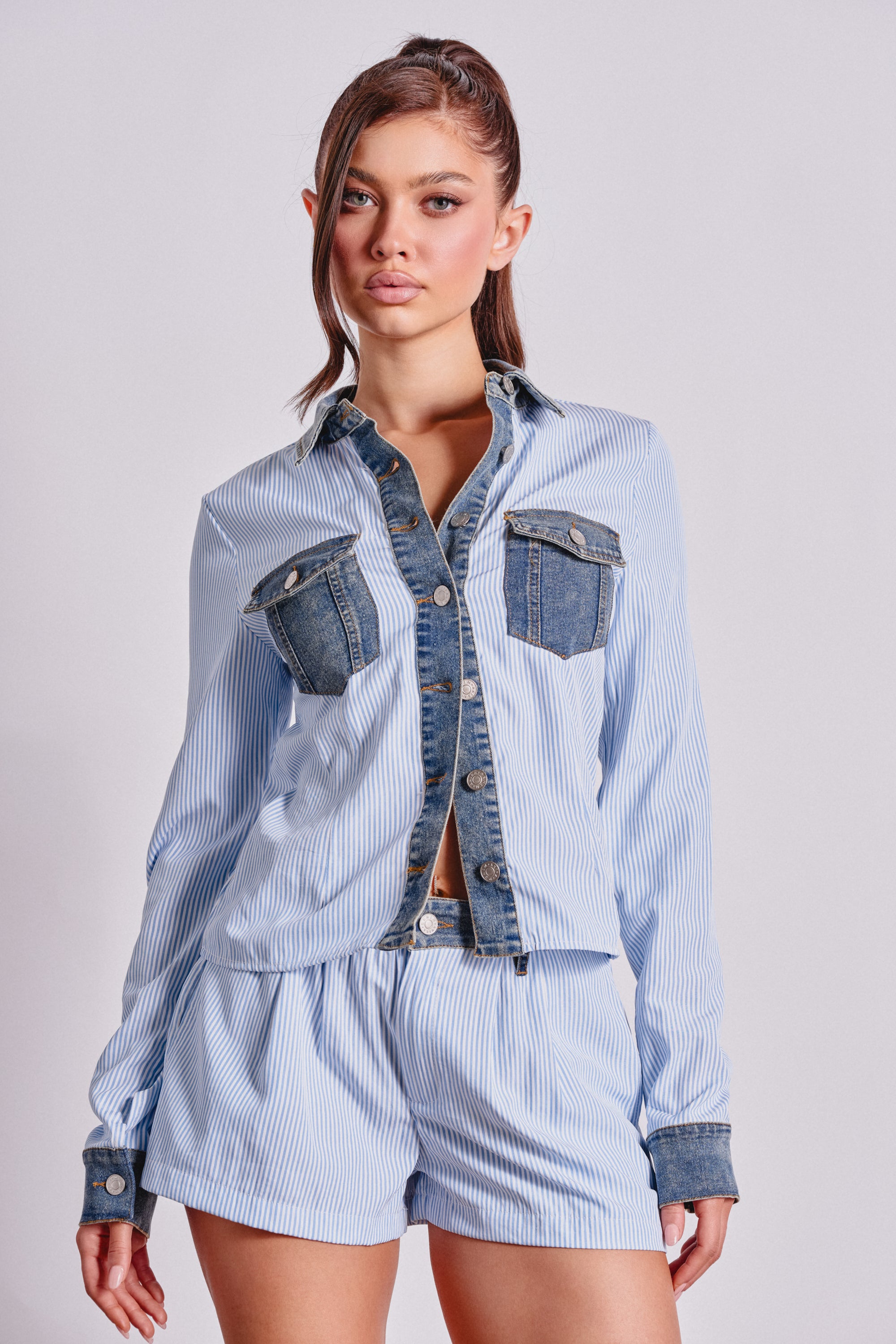 A woman models the EASY LOVER POPLIN BUTTON DOWN, a light blue and white pinstripe pajama-style top with denim trim on the collar, cuffs, and chest pockets, standing against a light gray background. 1
