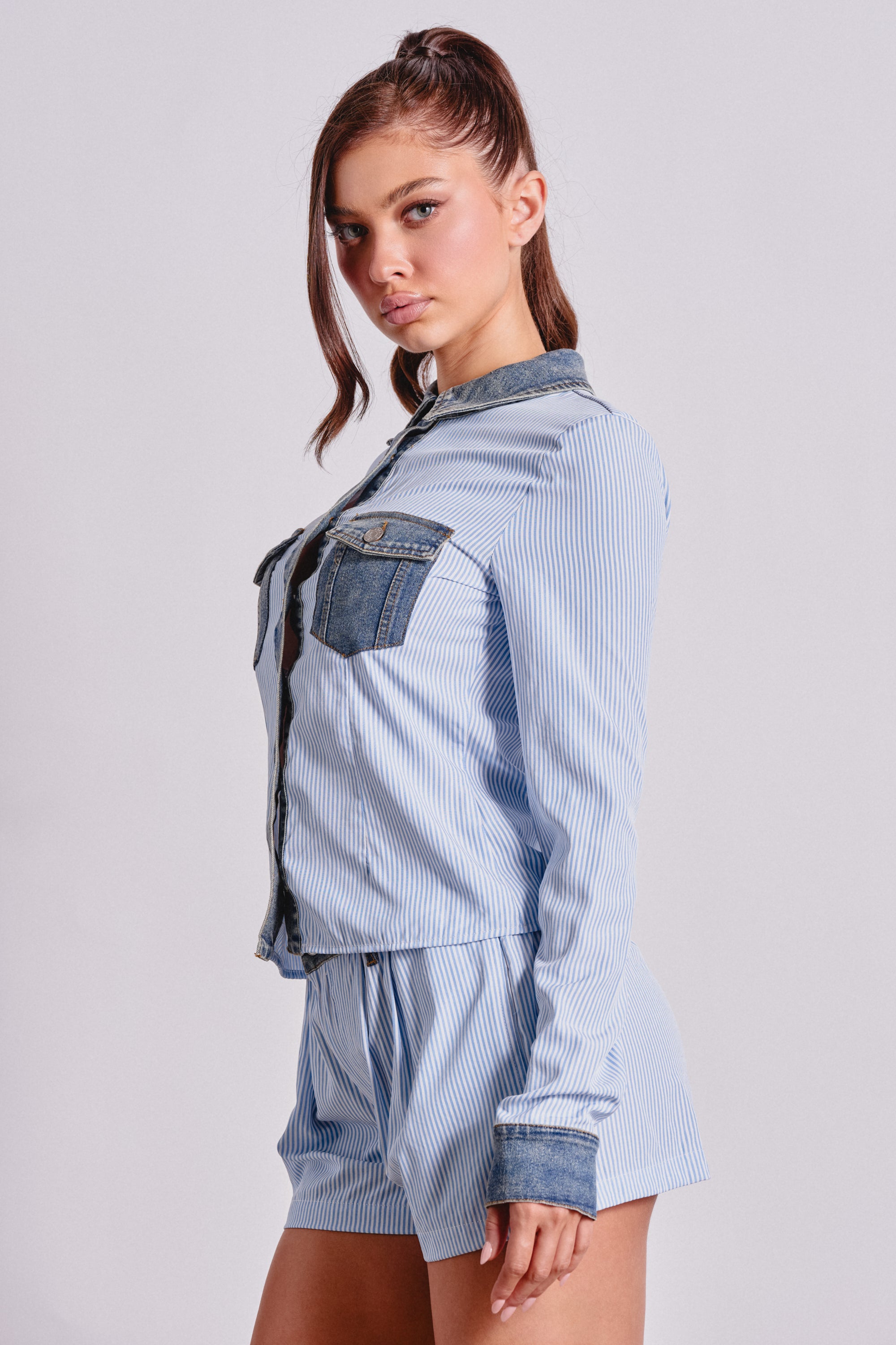 A young woman stands in profile against a plain light background, wearing the EASY LOVER POPLIN BUTTON DOWN—a pinstripe blouse with denim collar, cuffs, and pockets—paired with matching shorts. 2
