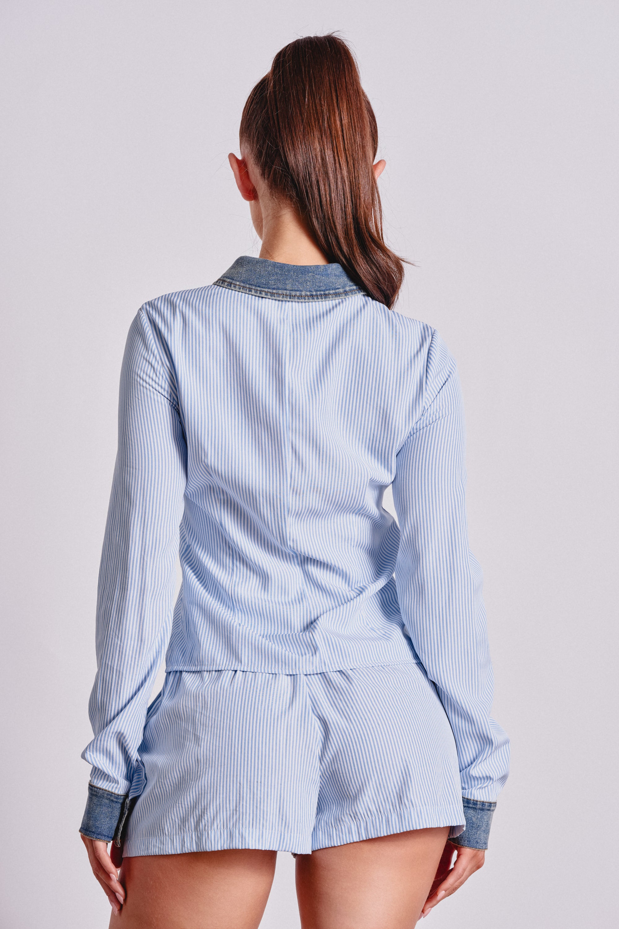 A person with long brown hair in a high ponytail is seen from behind, wearing the EASY LOVER POPLIN BUTTON DOWN in blue and white pinstripes with matching shorts, featuring dark cuffs and collar, against a plain background. 3