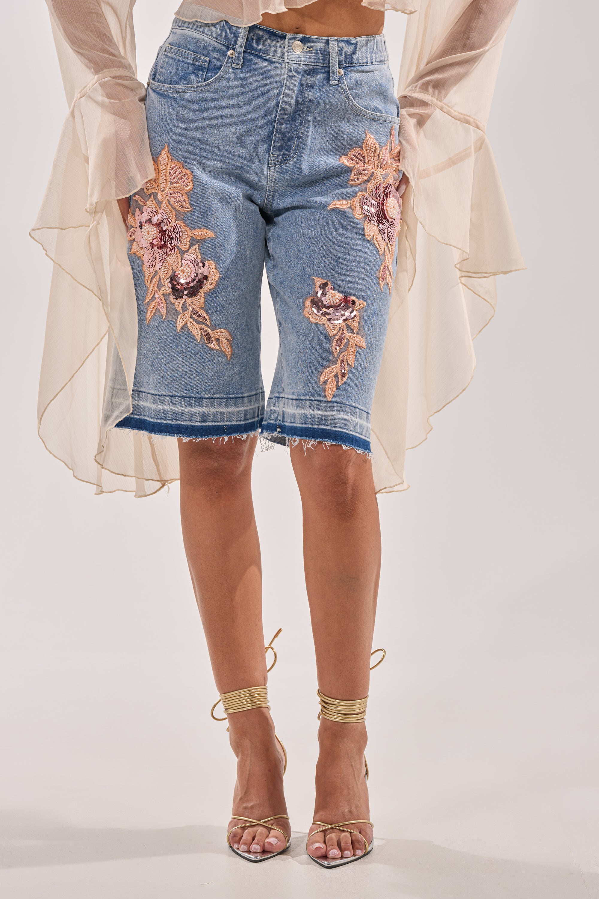 A person wears the TRY ME DENIM BERMUDA SHORT with a sheer, ruffled beige top and gold strappy high-heeled sandals. 0