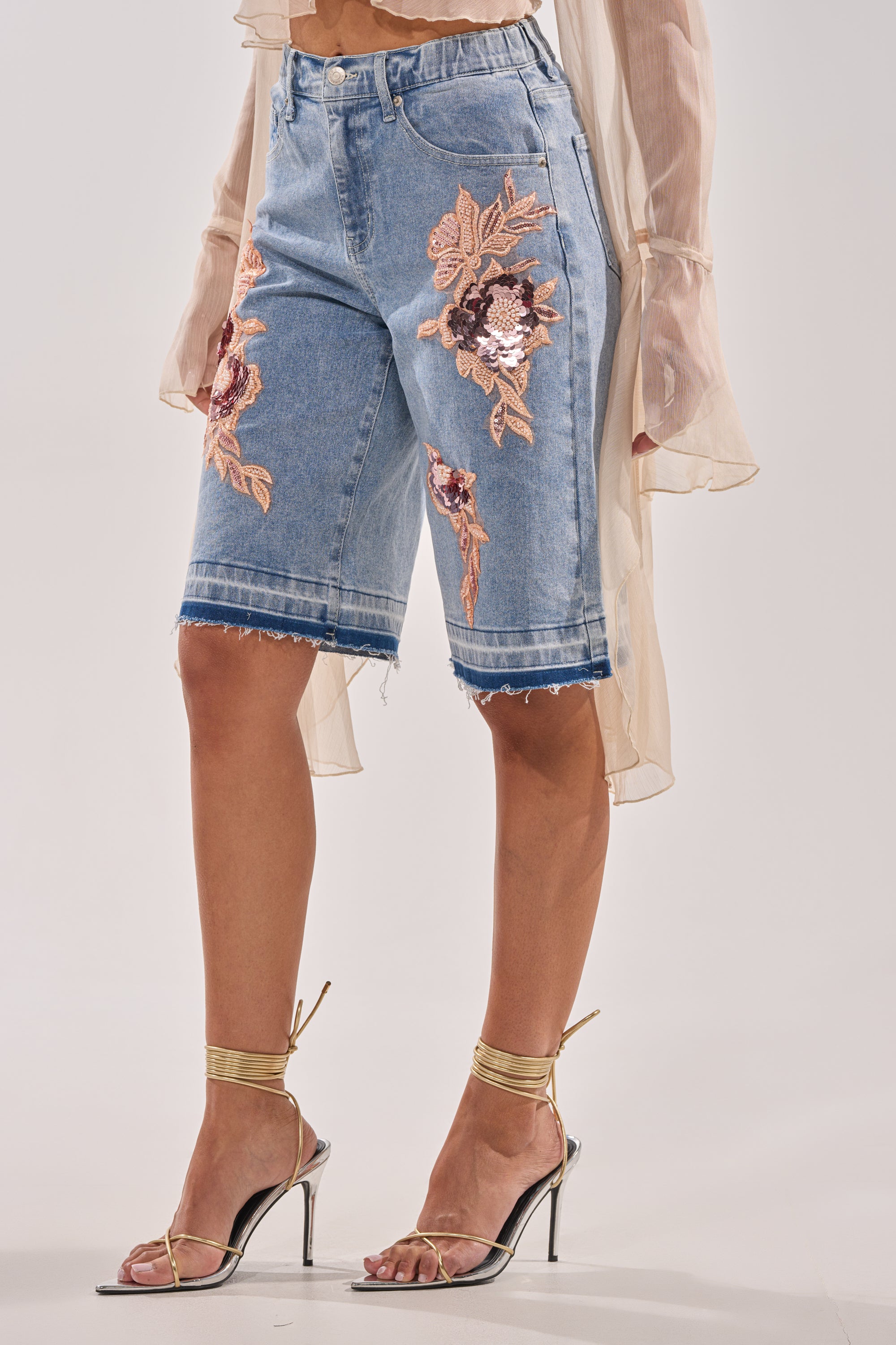 A person wears TRY ME DENIM BERMUDA SHORTS with pink floral embroidery, paired with a sheer long-sleeve top and metallic gold high-heeled sandals, showcasing a chic lower body ensemble. 2
