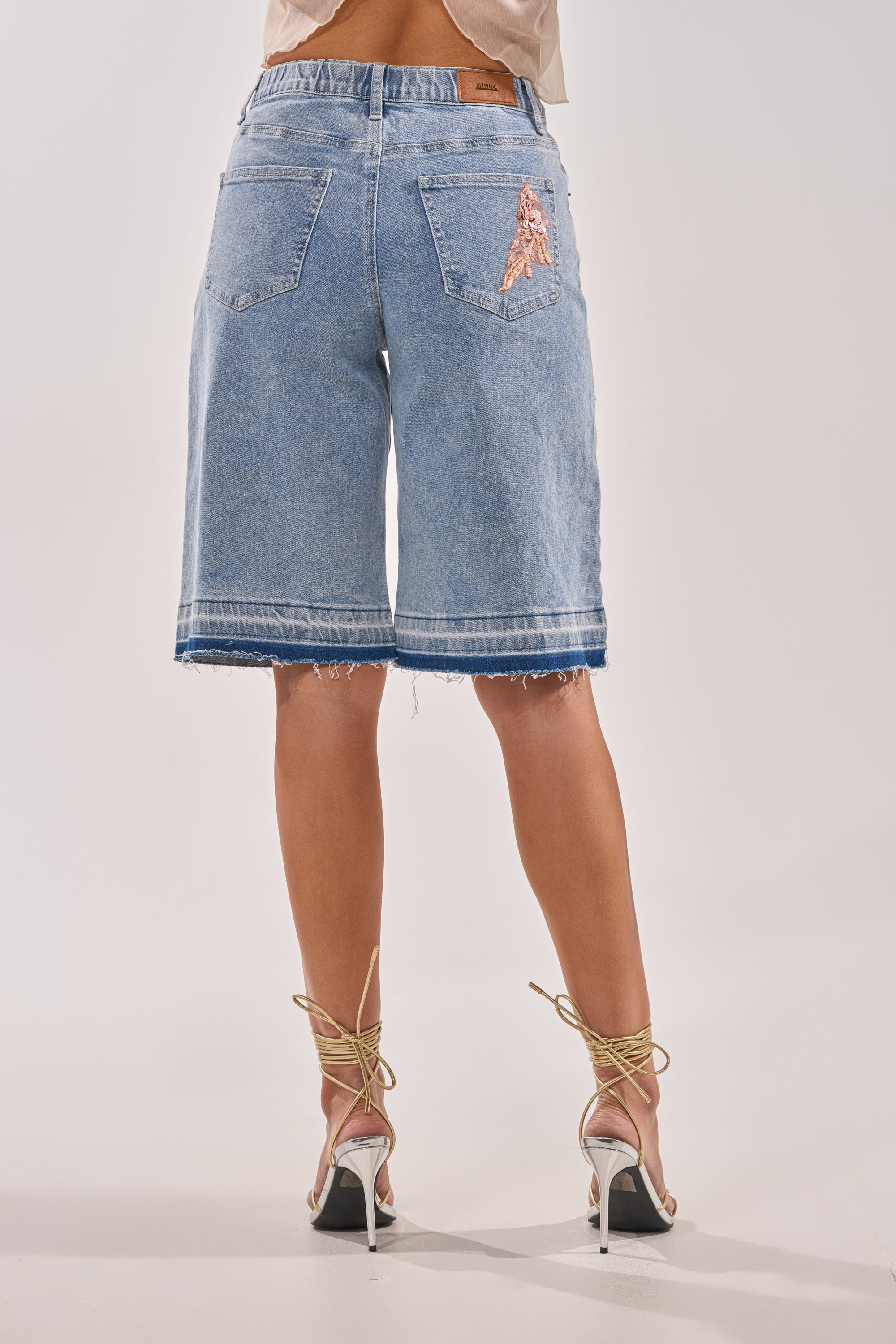 A person stands facing away, wearing the TRY ME DENIM BERMUDA SHORT—knee-length, light blue with frayed hems and two back pockets—paired with tan lace-up high-heeled sandals. 3