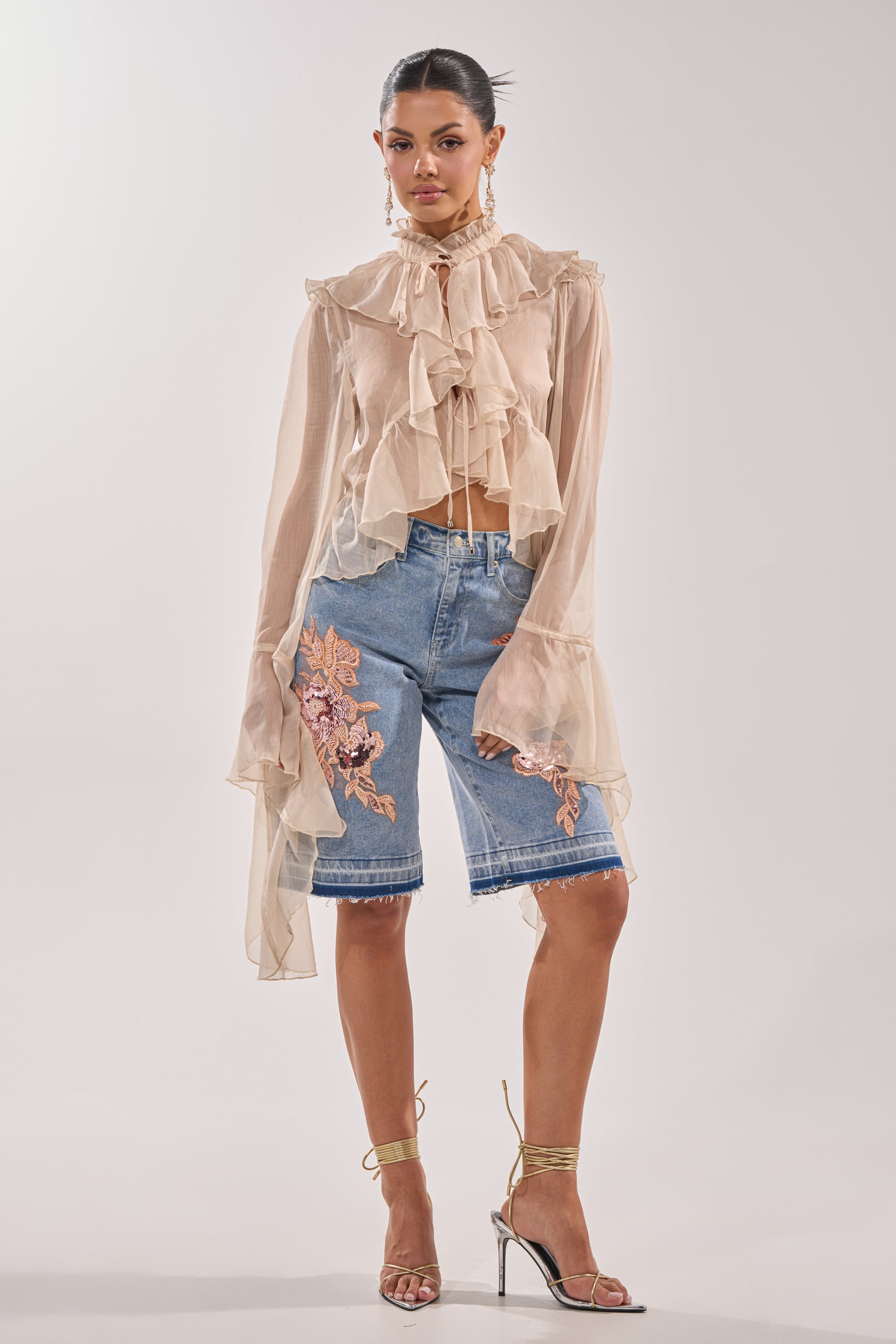 A woman stands against a plain background wearing a sheer beige ruffled blouse, gold strappy heels, drop earrings, and the TRY ME DENIM BERMUDA SHORT with pink floral embroidery. 4