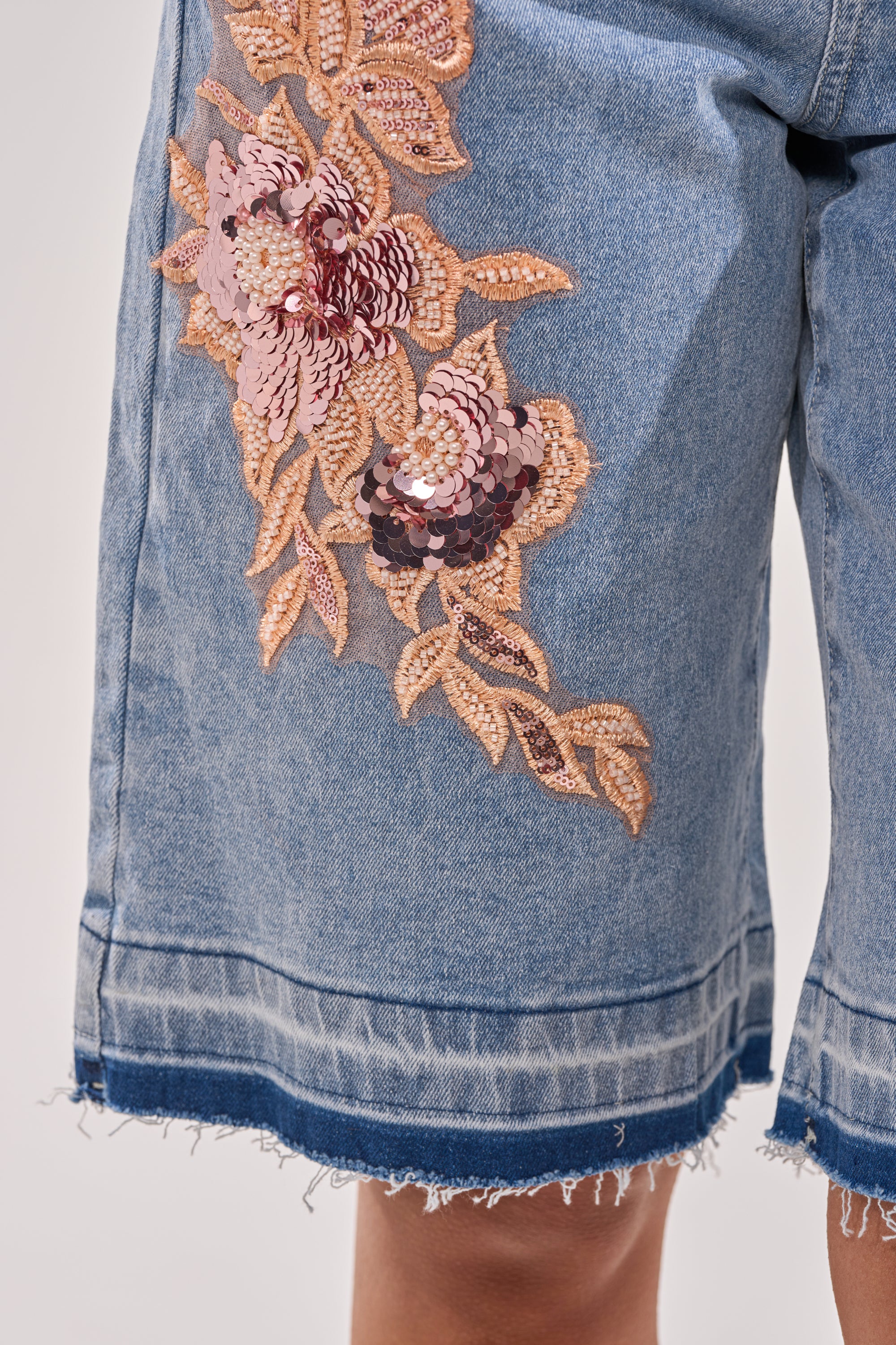 Close-up of the TRY ME DENIM BERMUDA SHORT, a high-rise short with frayed hems and a large pink and gold floral sequin applique on the left thigh. 5