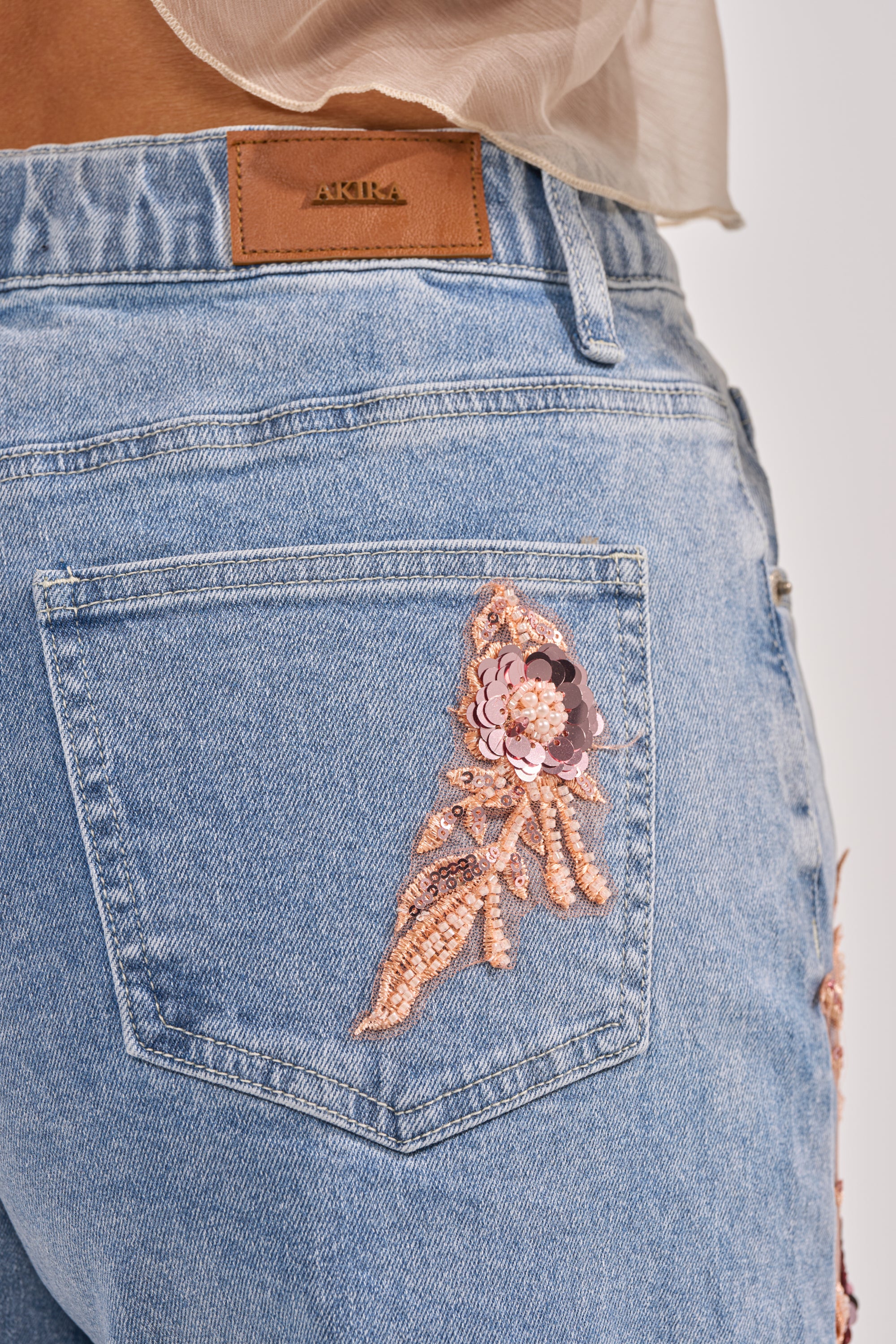 Close-up of the back pocket on the TRY ME DENIM BERMUDA SHORT in light blue with pink floral embroidery and gold accents. A brown leather "AKIRA" patch is sewn above the pocket, highlighting the high-rise style. 6