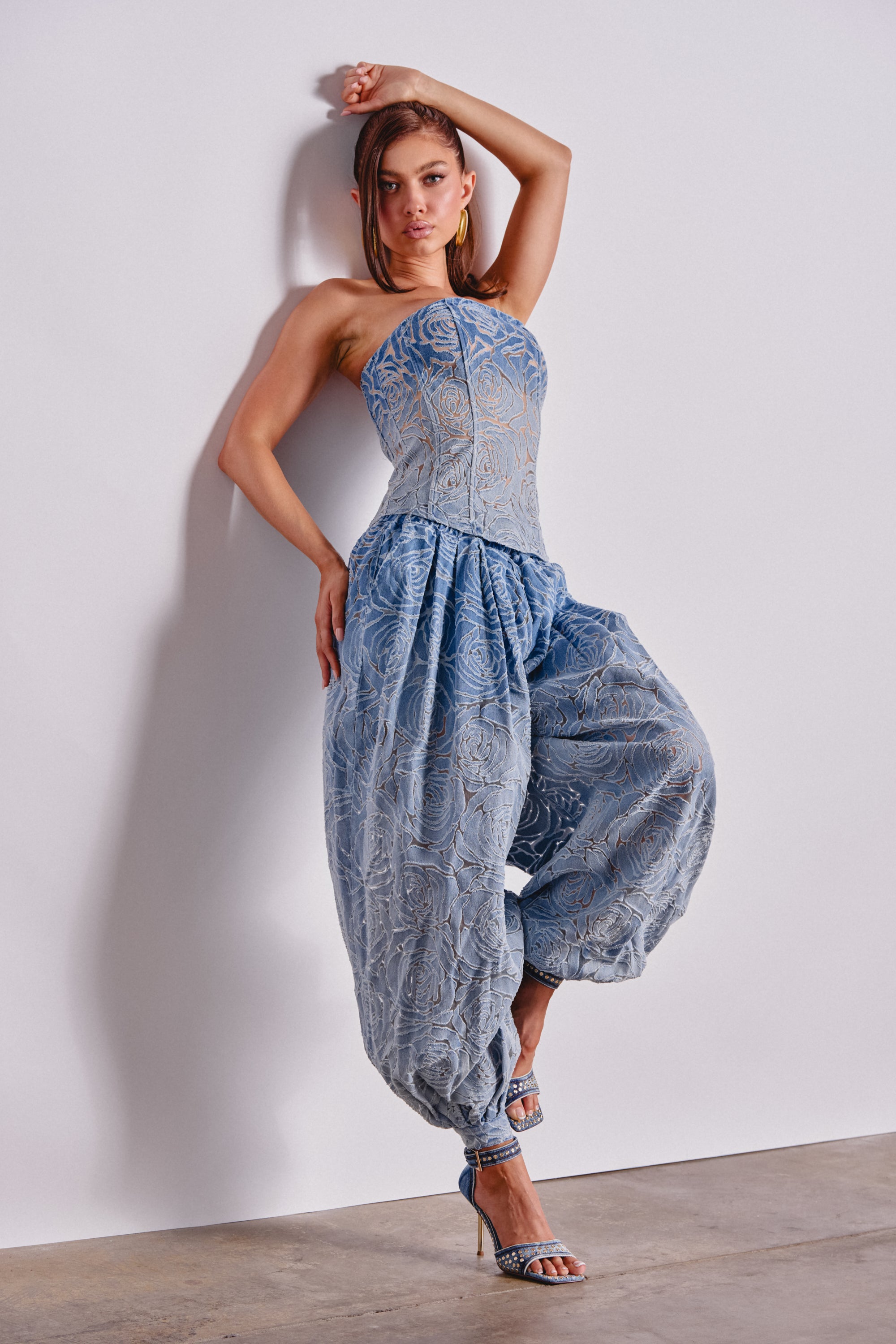 A woman in a blue rose-patterned corset and DENIM FOR DAYS BALLOON PANT leans against a white wall with one arm raised, wearing strappy heels. 0