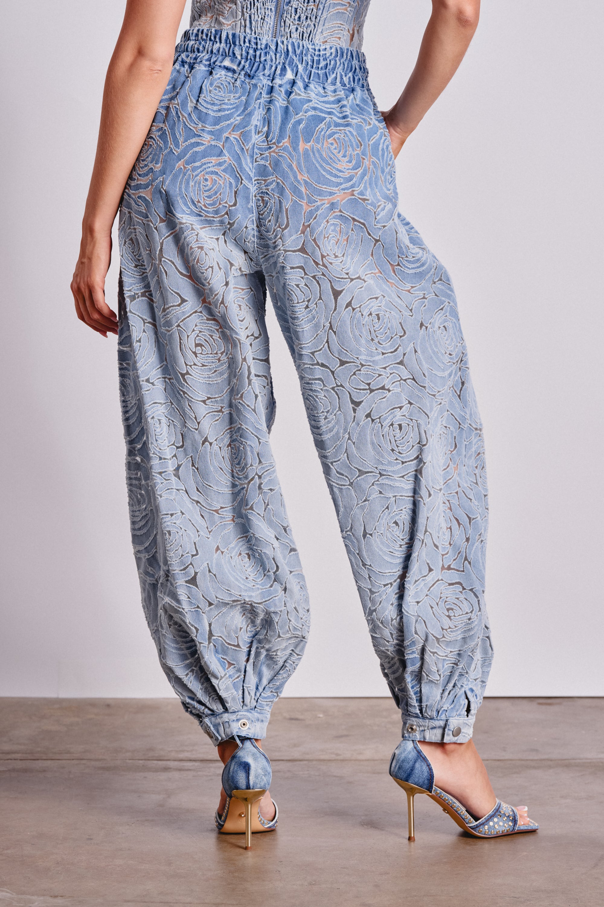 A person stands on a concrete floor wearing the DENIM FOR DAYS BALLOON PANT—light blue, high-rise sheer lace pants with a floral pattern and cinched ankles—paired with blue high-heeled sandals. 3