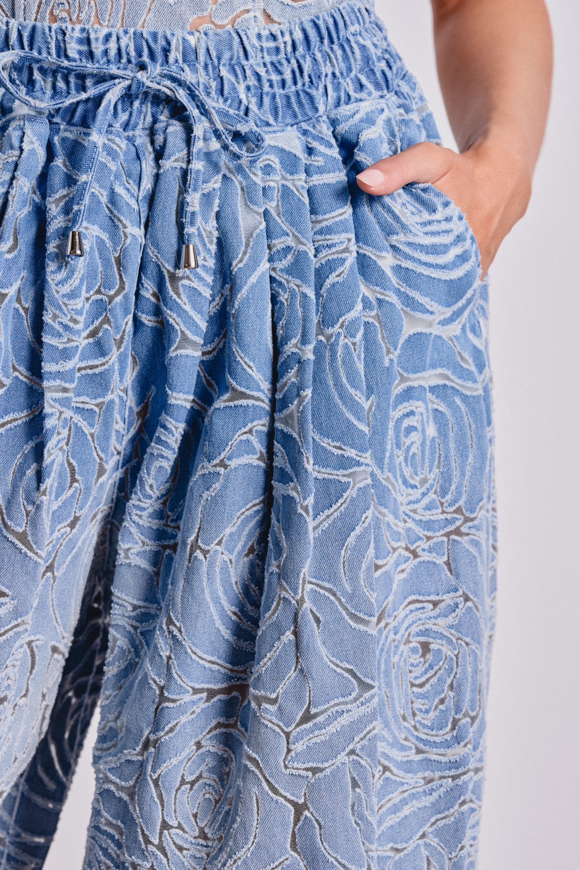 Close-up of a person wearing the DENIM FOR DAYS BALLOON PANT in light blue, featuring a textured floral pattern, elastic waistband with drawstrings, and their right hand in the pocket. 4