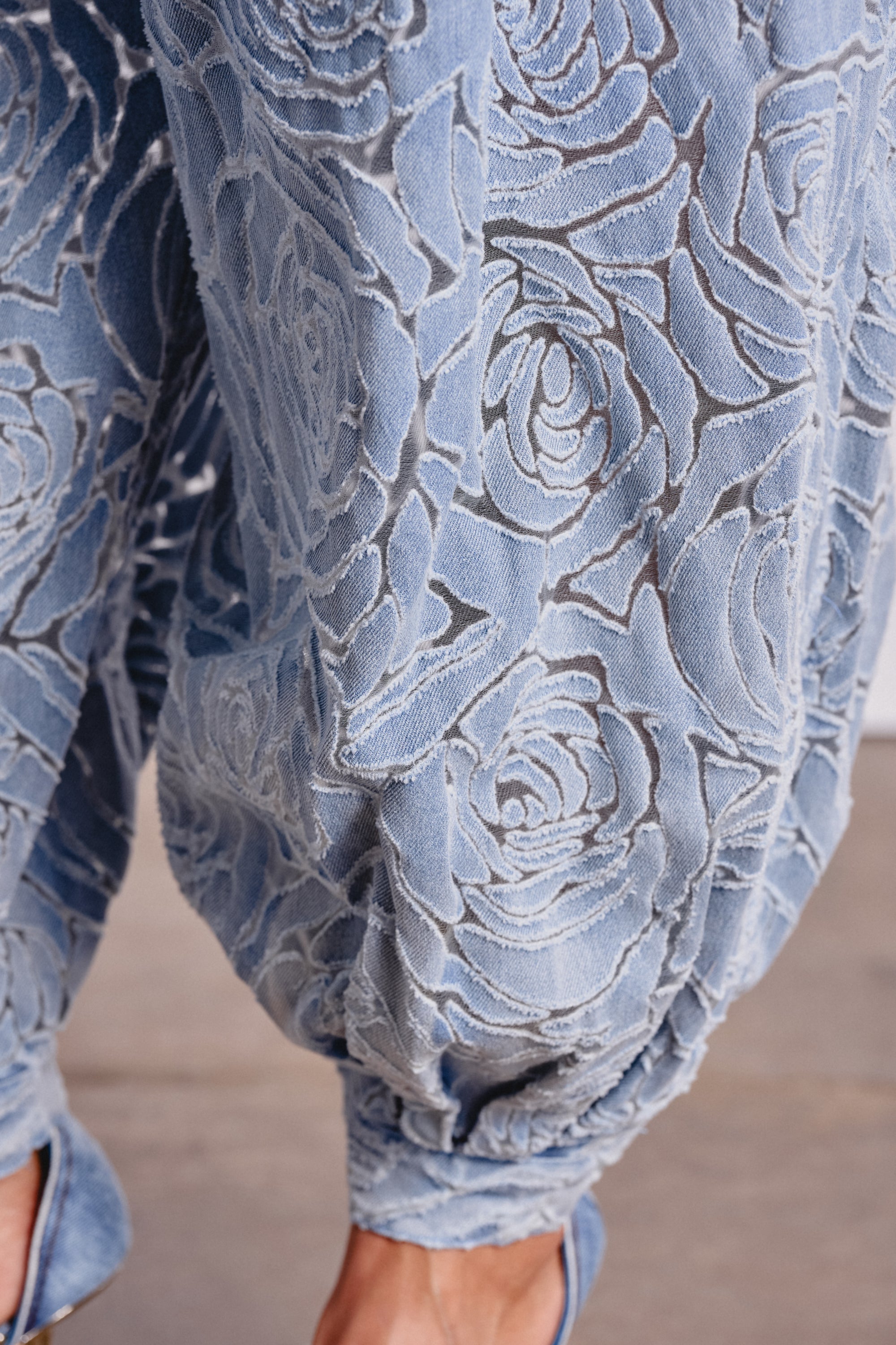 Close-up of the Denim For Days Balloon Pant in light blue denim, featuring an embossed floral rose pattern, high-rise fit, gathered ankle cuffs, and a peek at matching blue heels. 5